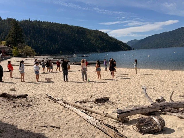 Group of people on a sandy beach near a lake, backed by forested hills under a partly cloudy sky.