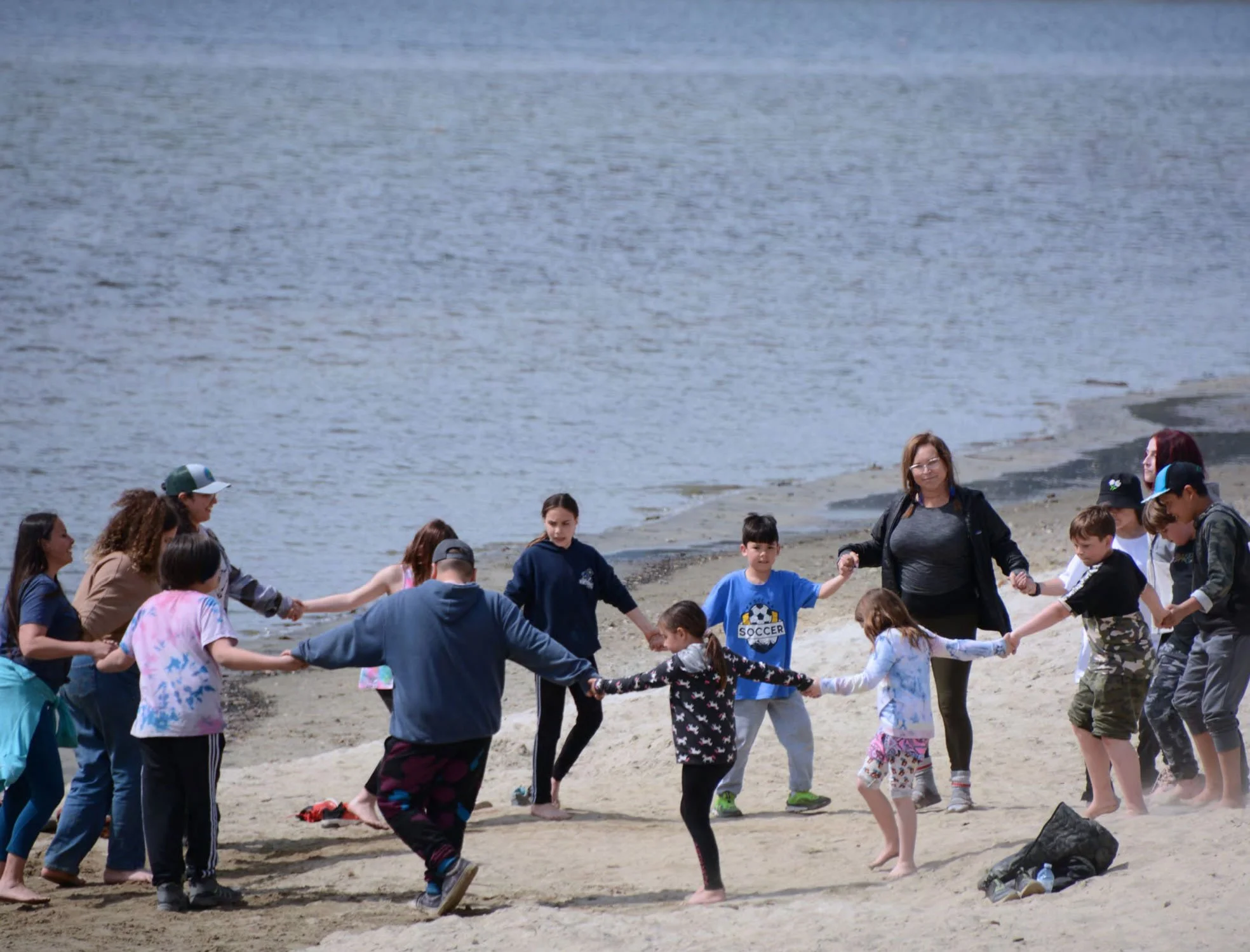 A group of children and adults holding hands and forming a circle on a beach near the water, engaging in a group activity.