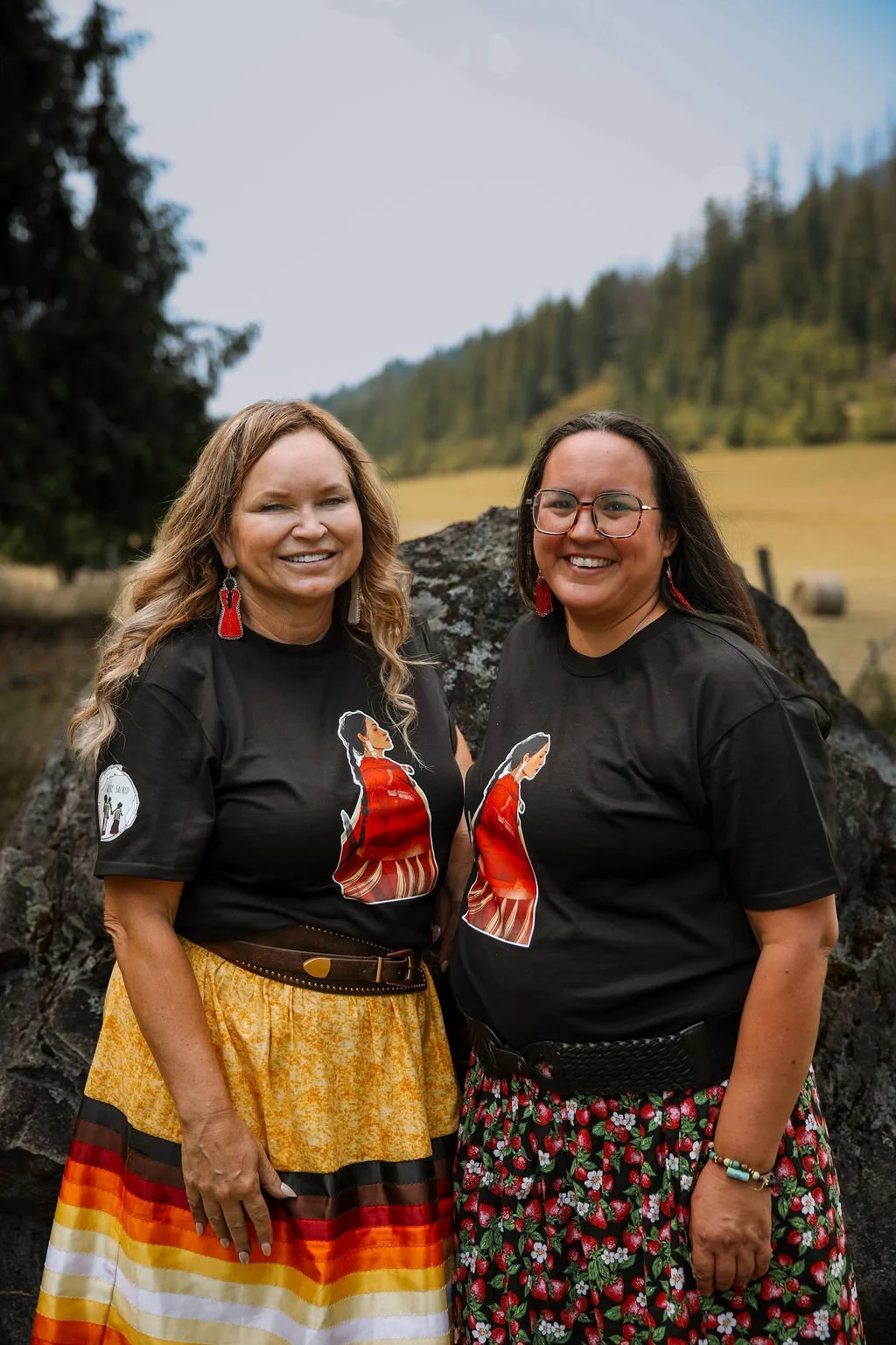 Two women standing outdoors in front of a large rock, wearing matching black T-shirts with a woman in a red dress and a long skirt design, smiling at the camera, with a forested hillside and cloudy sky in the background.