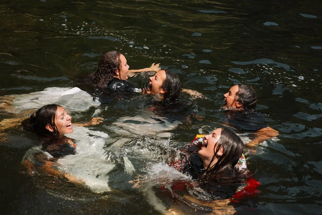 Five women are swimming and playing in a body of water, splashing and smiling.