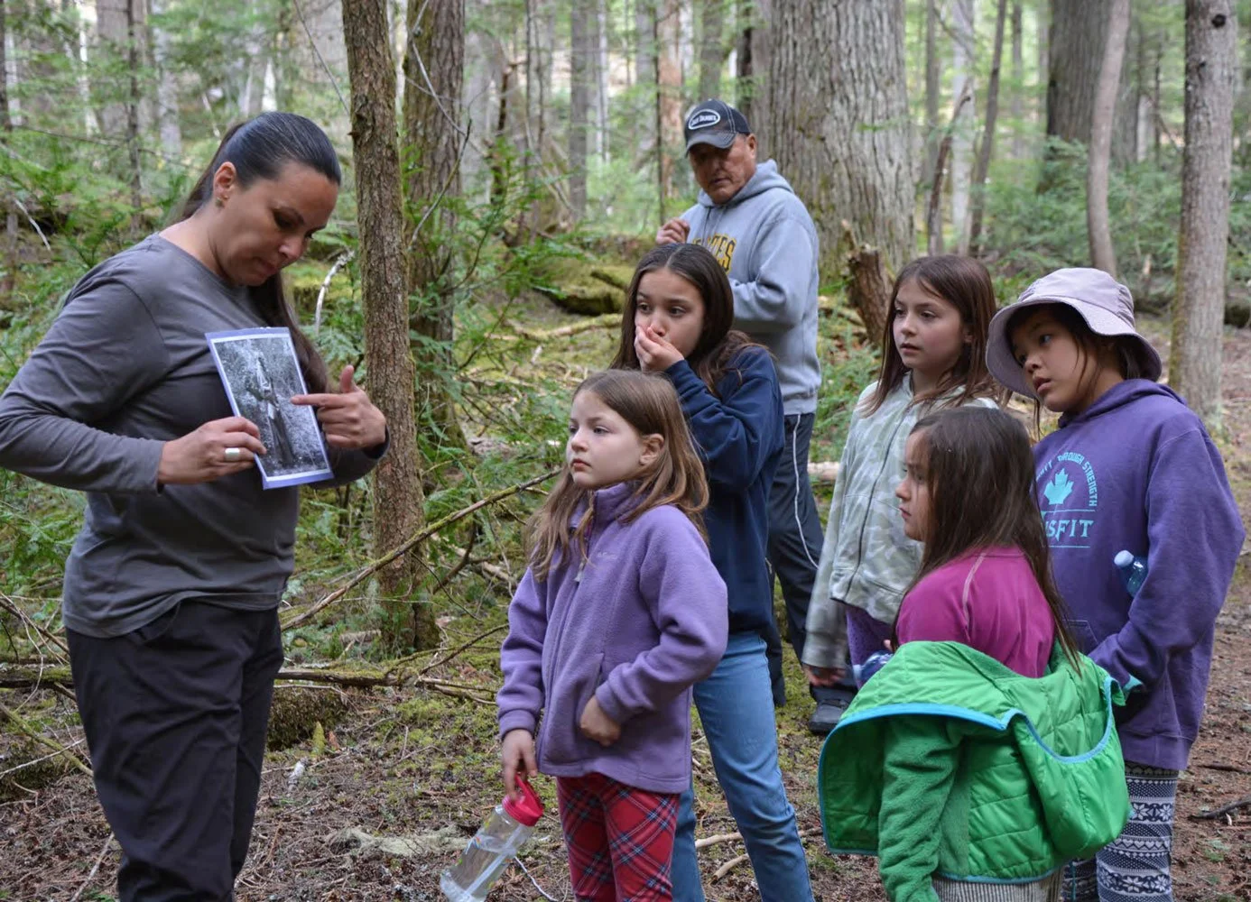 A woman leading a nature educational activity with six children in a forest, holding a black-and-white photograph.