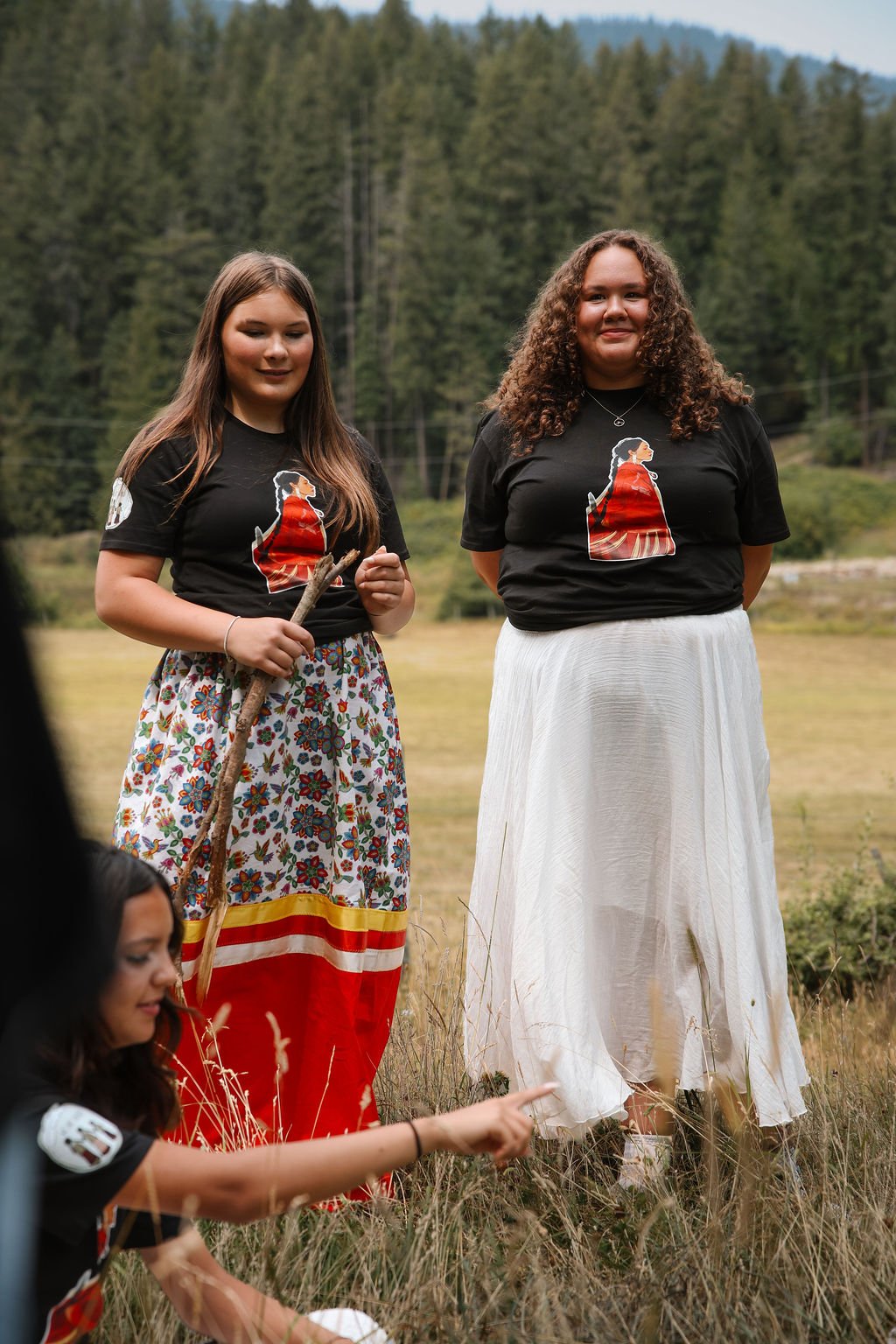 Two women standing outdoors in a grassy field with a forested hillside behind them. They are wearing matching black T-shirts with a graphic of a woman in a red dress. One woman has curly hair and the other has straight hair. A third person is partially visible in the foreground, reaching toward the ground.