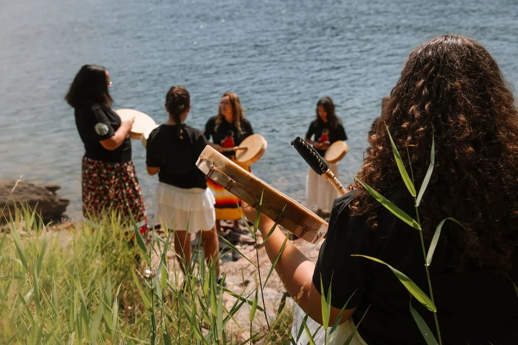 A group of women drumming by the water, with one woman playing a small handheld percussion instrument and others holding drums, near a rocky shoreline with grass in the foreground.