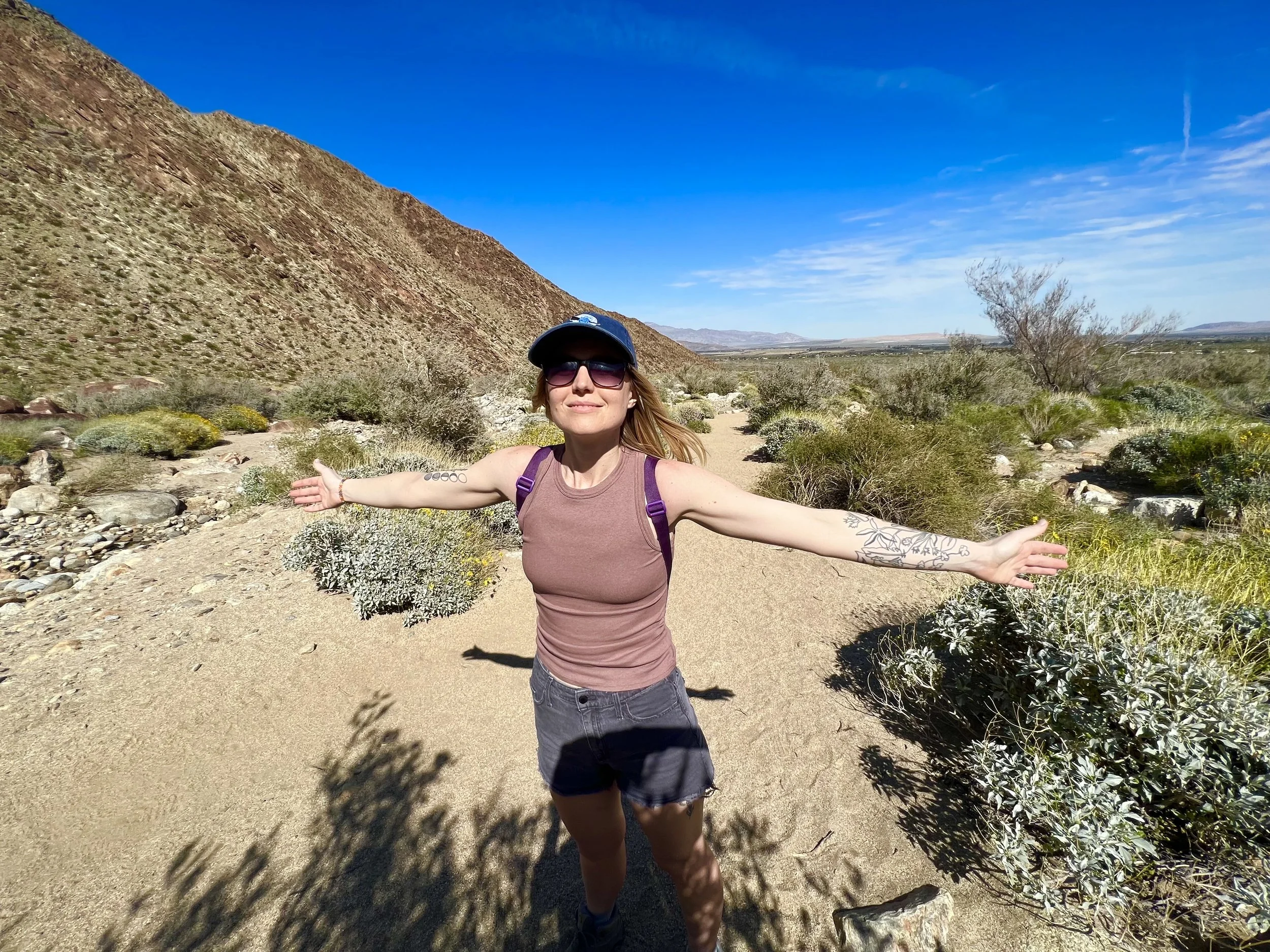 Lisa Williams stands with outstretched arms on a desert trail with desert plants and mountains in the background under a blue sky.