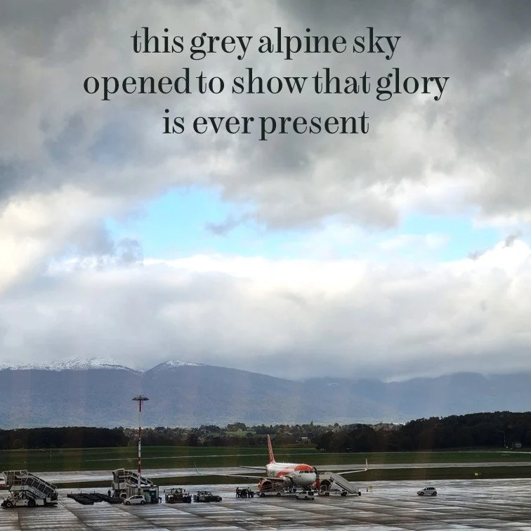 An airplane parked on the tarmac at an airport with stairs and service vehicles around. In the background, there are mountains partly covered with snow under a mostly grey alpine sky with some patches of blue.