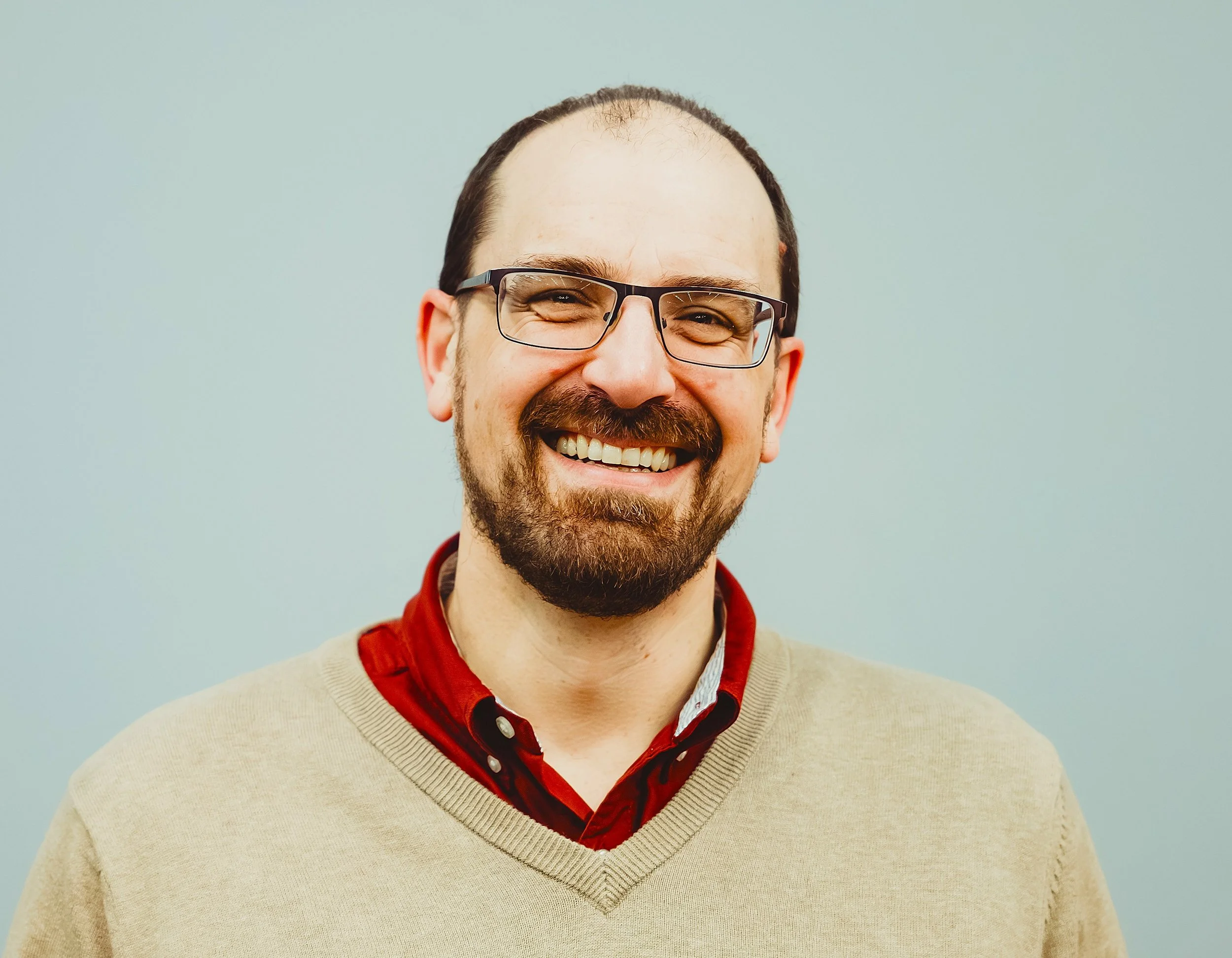 Headshot of Dr. Steve Jones, smiling with glasses, beard, wearing a beige sweater over a red collared shirt..