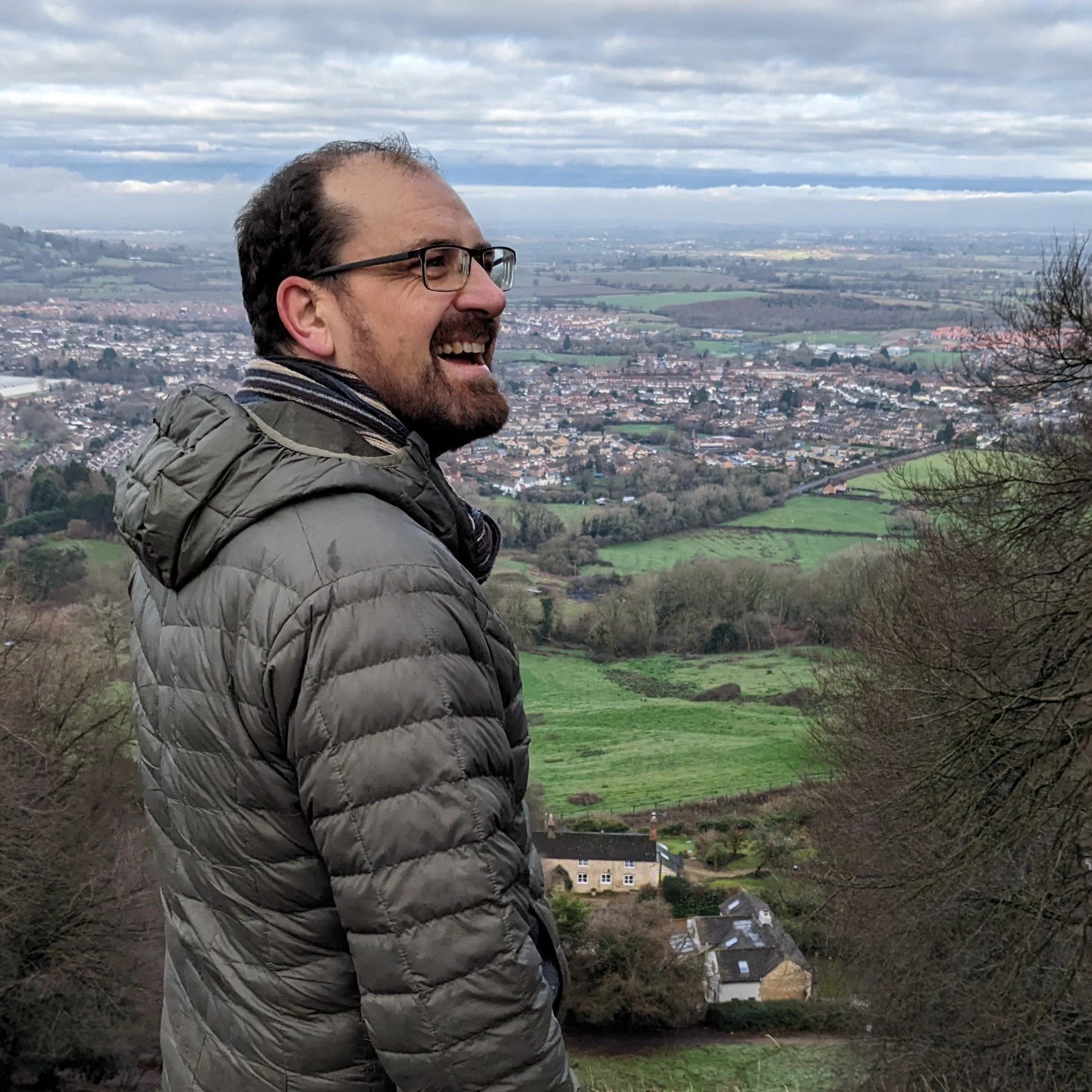 Picture of Dr. Steve Jones, wearing glasses and a puffy jacket, standing outdoors with a panoramic view of a the Severn Valley in the background under a cloudy sky.