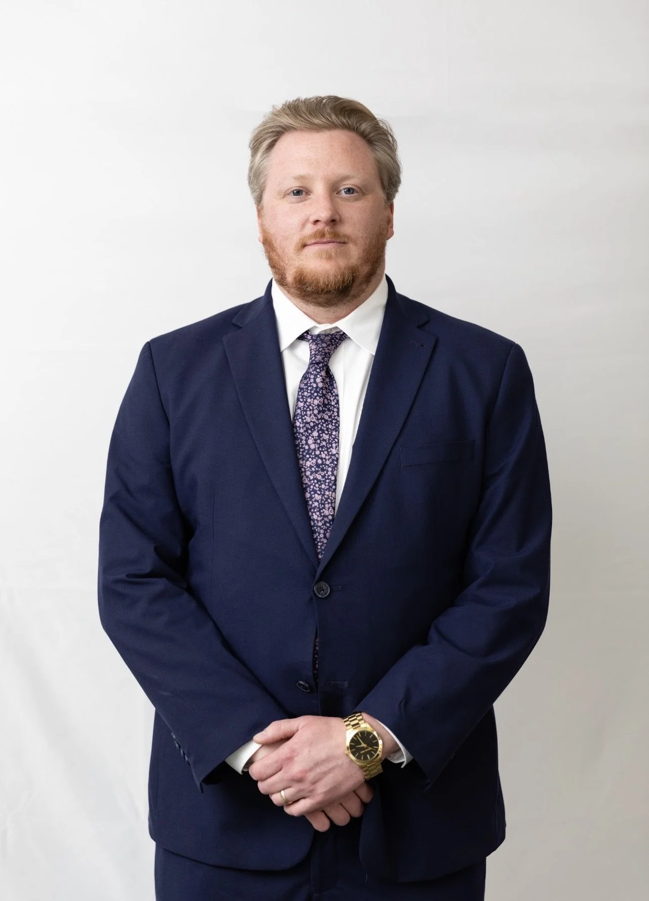 A man with light skin, red hair, and a beard wearing a navy blue suit, white shirt, floral purple tie, gold wristwatch, and a wedding band, standing against a plain white background.