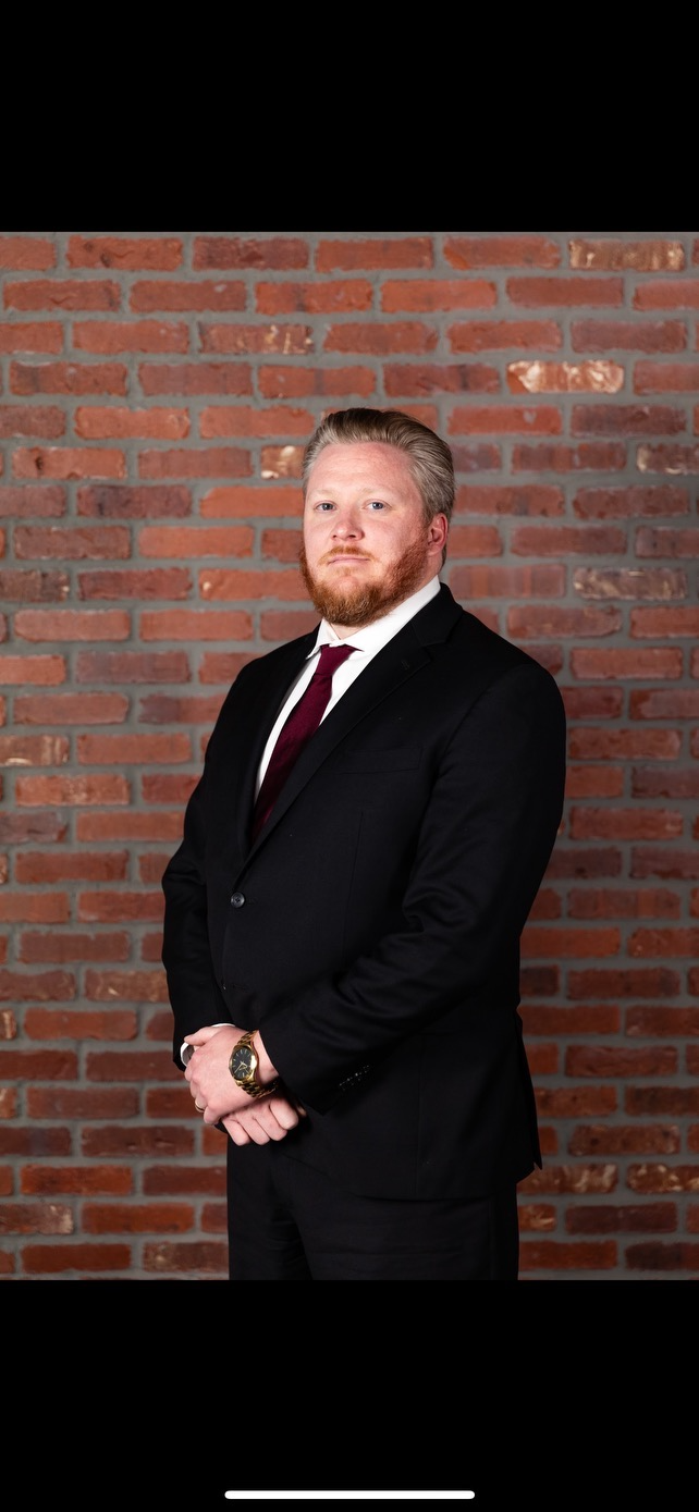 A man in a black suit with a white shirt and maroon tie standing in front of a brick wall, looking at the camera with a serious expression.