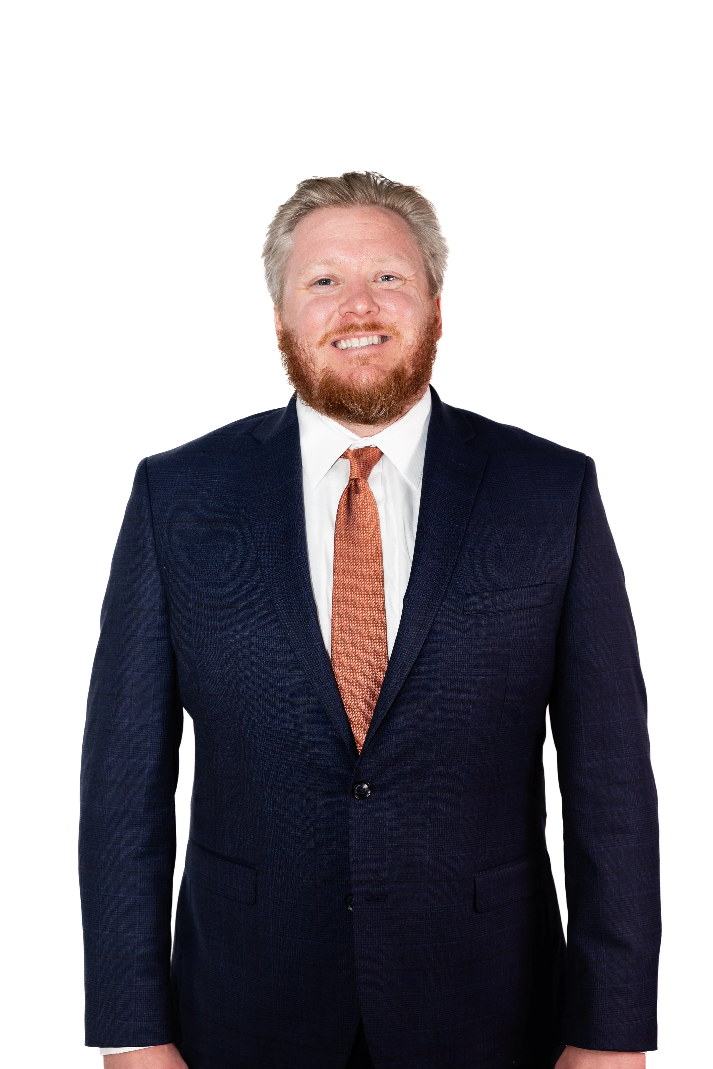 A man with a red beard and grey hair wearing a navy suit, white shirt, and orange tie, smiling against a white background.