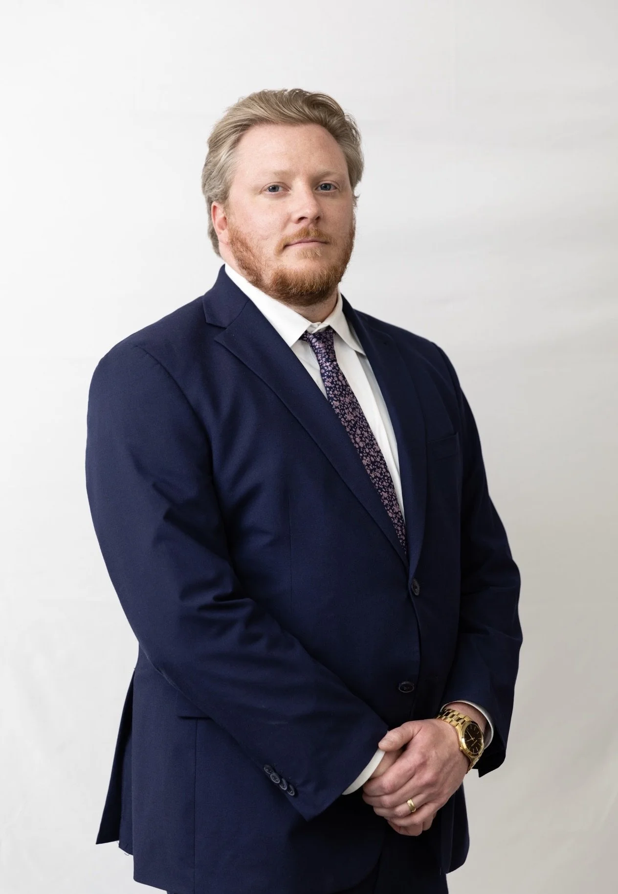 A man wearing a dark blue suit with a white shirt and patterned tie, standing against a plain white background. He has light-colored hair, a red beard, and is wearing a gold watch and a wedding band.