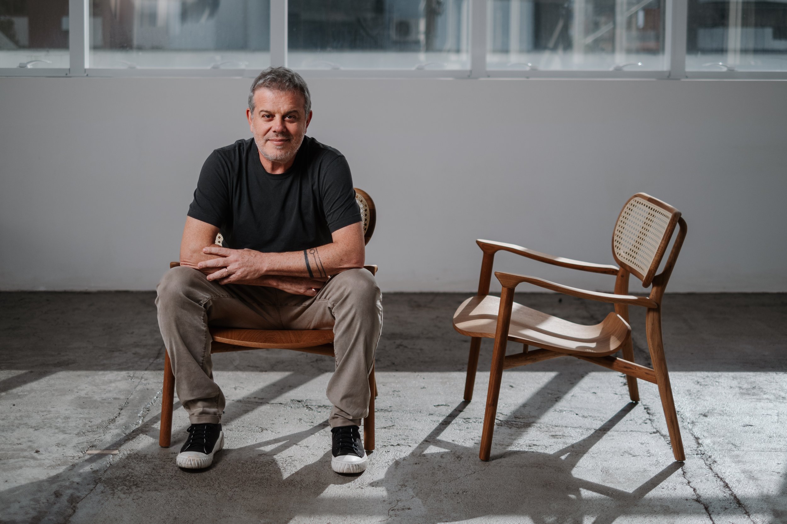Homem sentado em uma cadeira de madeira, usando camiseta preta, calça bege, tênis preto e branco, com um sorriso moderado, em um ambiente com parede cinza e uma janela ao fundo. Ao lado dele, uma cadeira de madeira com encosto de coxim de palhinha.
