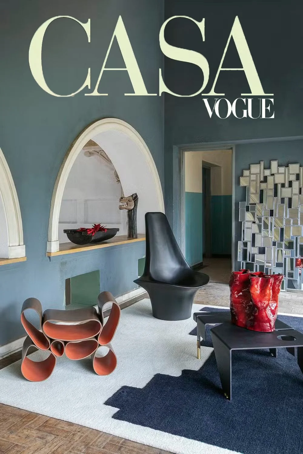 Interior de uma sala decorada com móveis modernos, incluindo uma cadeira preta de design único, uma mesa baixa preta com vasos vermelhos, uma parede azul com arcos brancos, e uma parede de espelhos em formato de cubos, com uma porta ao fundo.