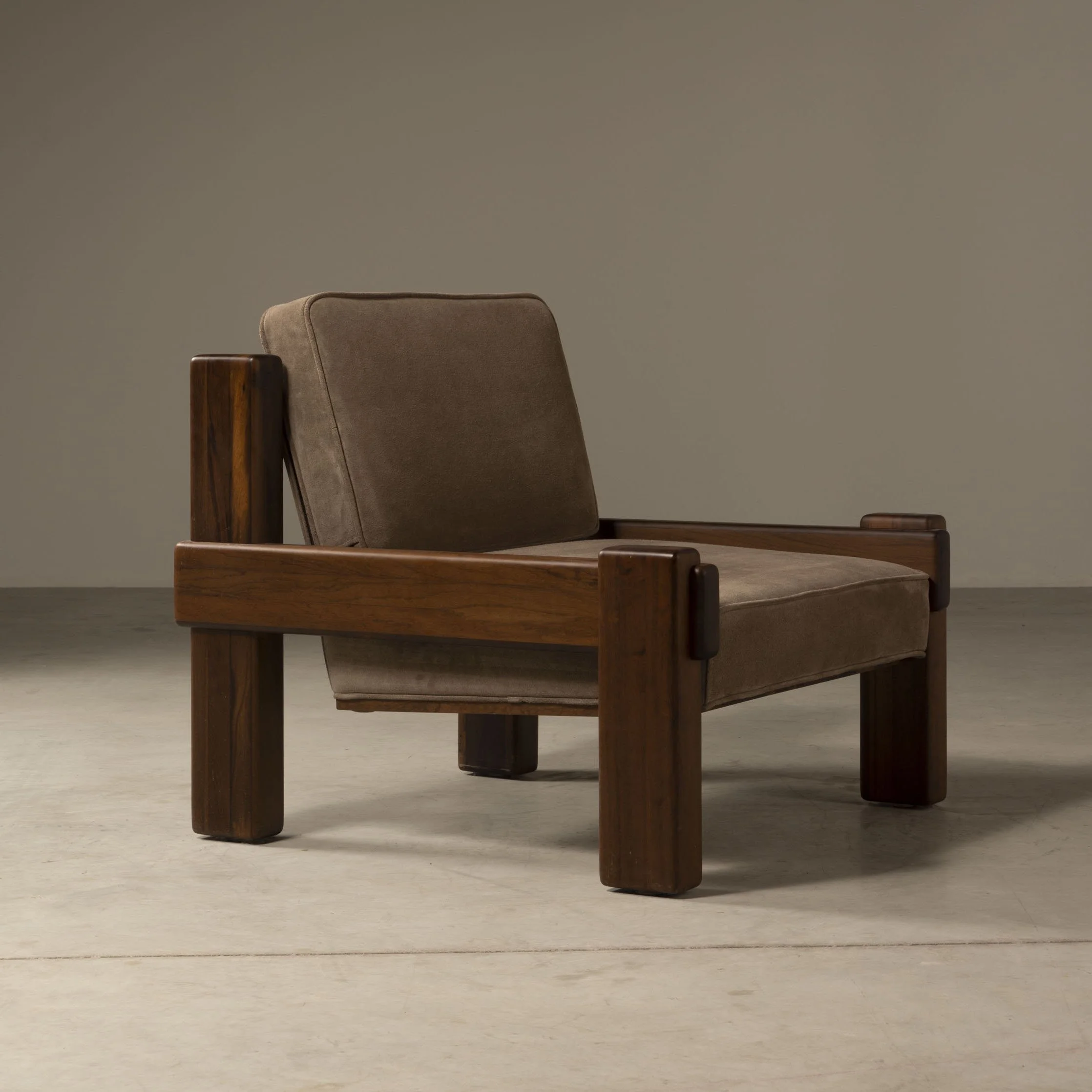 Cadeira de madeira com almofada bege na parte de trás e mesa ao lado, ambiente com parede cinza claro e piso de concreto.
