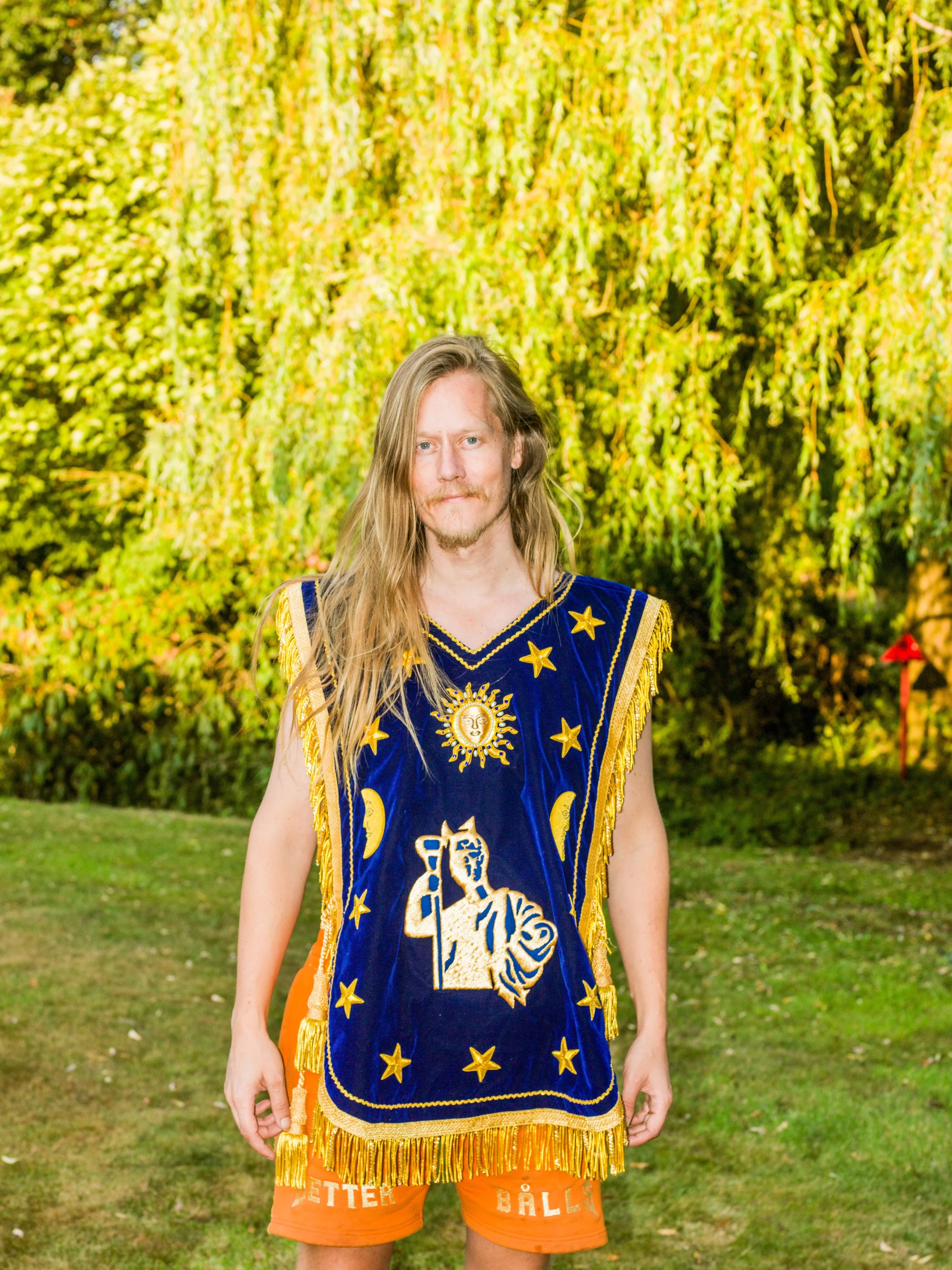 Jakob Vanderblank wearing a custom hand-embroidered BETTER BÅLLS garment in blue and gold, standing in a lush garden.