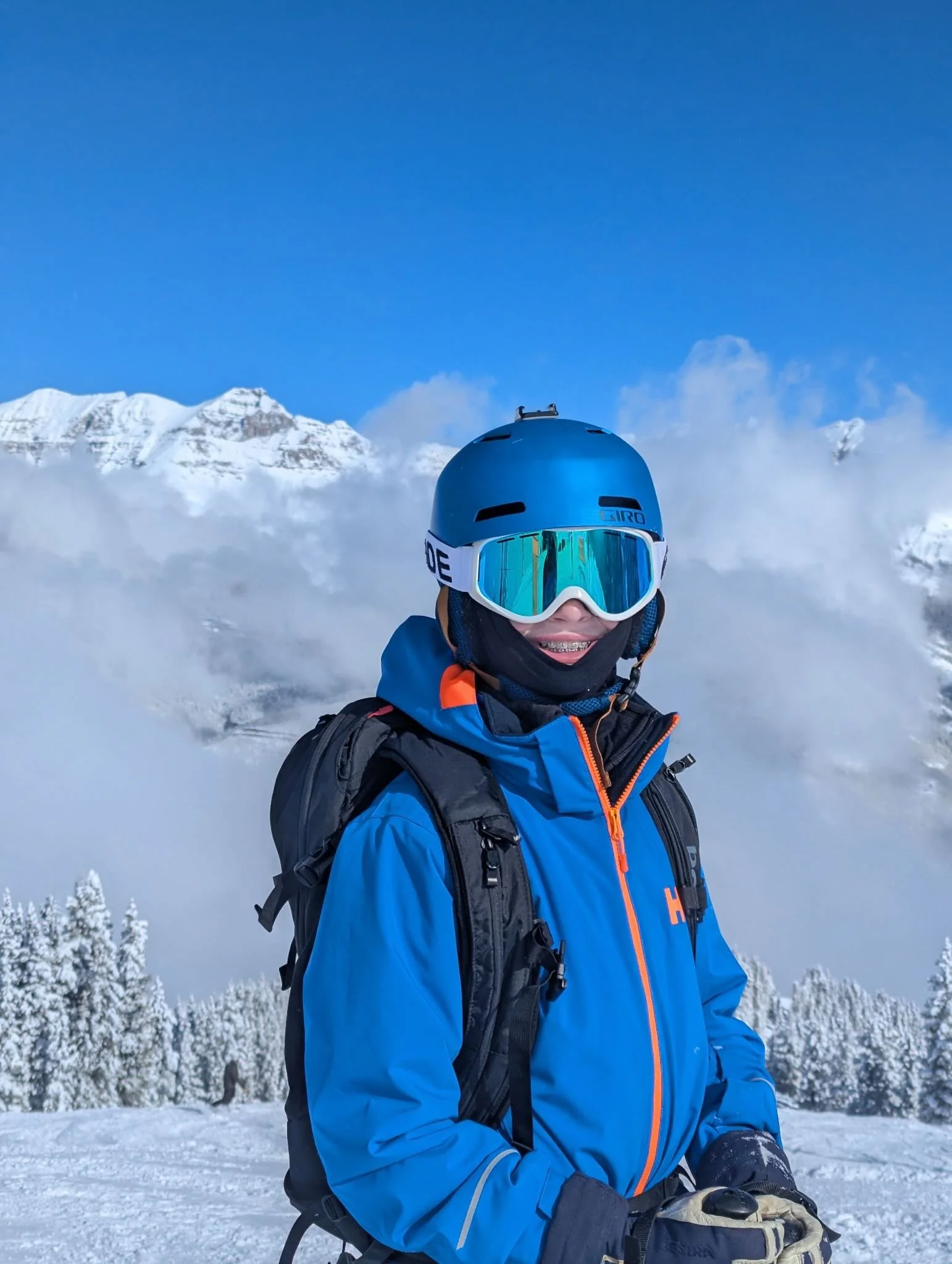 A person wearing a blue winter jacket, black gloves, and a helmet with goggles takes a selfie in a snowy mountain landscape with snow-covered peaks and frost-covered trees in the background.