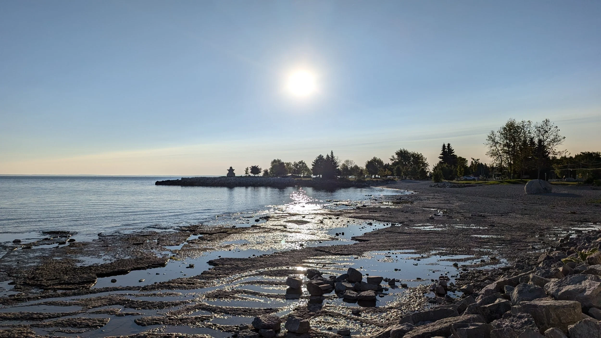 A tranquil shoreline scene with a sandy and rocky beach, calm water, and a clear sky with the sun shining overhead. There are trees and a small structure on a distant landform across the water.