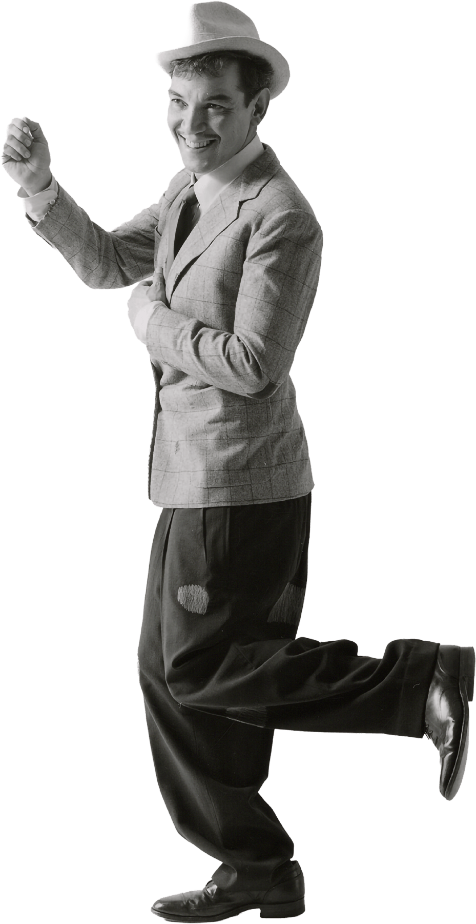Cantinflas con sombrero, saco a cuadros, camisa blanca, corbata y pantalones anchos con parches, ajustados con zapatos formales. Practicando un paso de baile y sonriendo.
