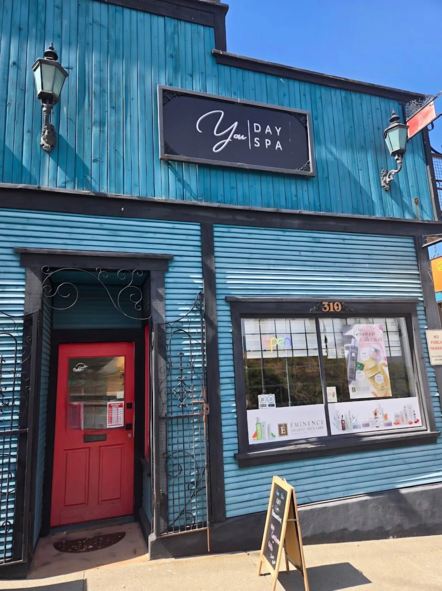 The exterior of a building with blue vertical siding, a red door, a large window, and a sign reading 'Your Day Spa' on the upper part of the building. There is a blackboard sign on the sidewalk and two vintage-style wall-mounted lamps on either side of the sign.