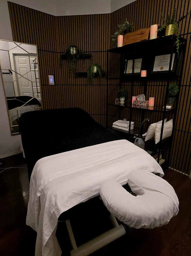 Massage table in a cozy room with wooden-paneled walls, decorated with plants, candles, and shelves holding framed certificates and towels.