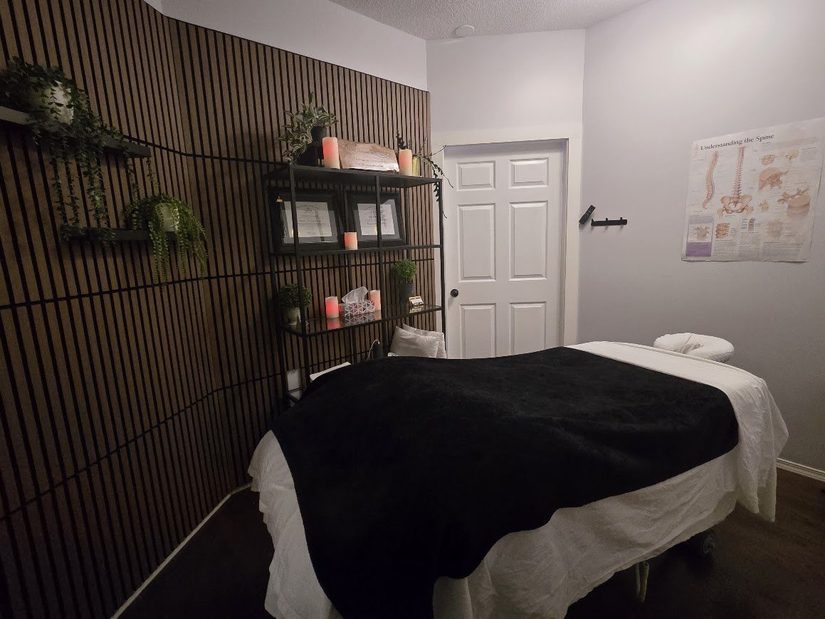 A massage therapy room with a massage table covered in white linens and a black blanket, a wooden slat accent wall with decorative shelves holding plants, candles, and framed photos, a closed white door, a poster about the spine on the wall, and a pillow and rolled towel on the table.