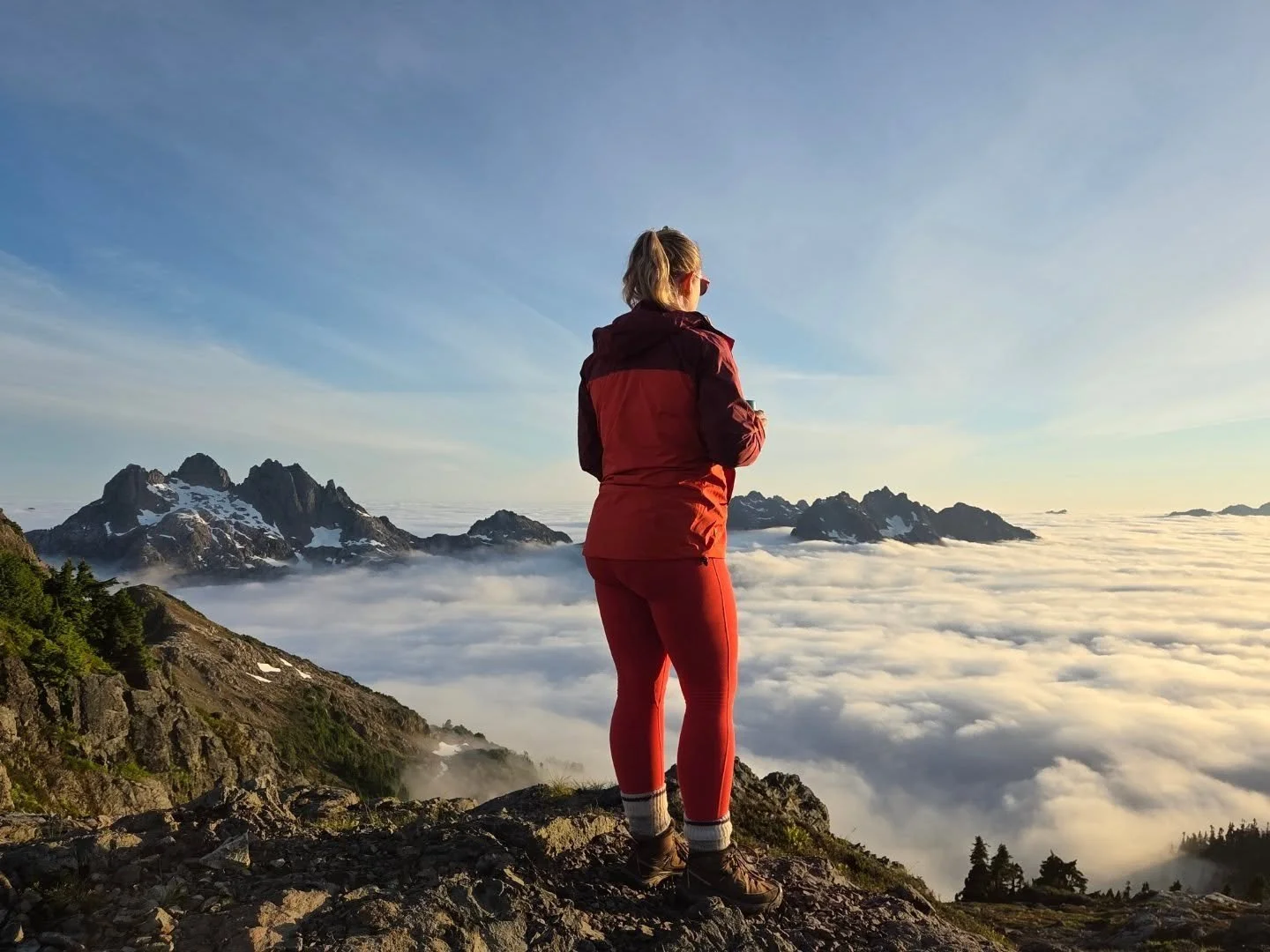A woman dressed in outdoor gear standing on a rocky mountain overlooking clouds and distant snow-capped peaks during sunrise or sunset.