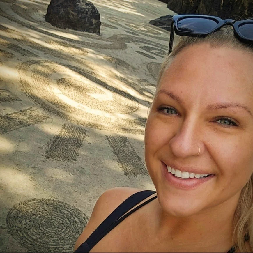 Woman with sunglasses on her head taking a selfie on the beach with tire tracks and rocks in the background.