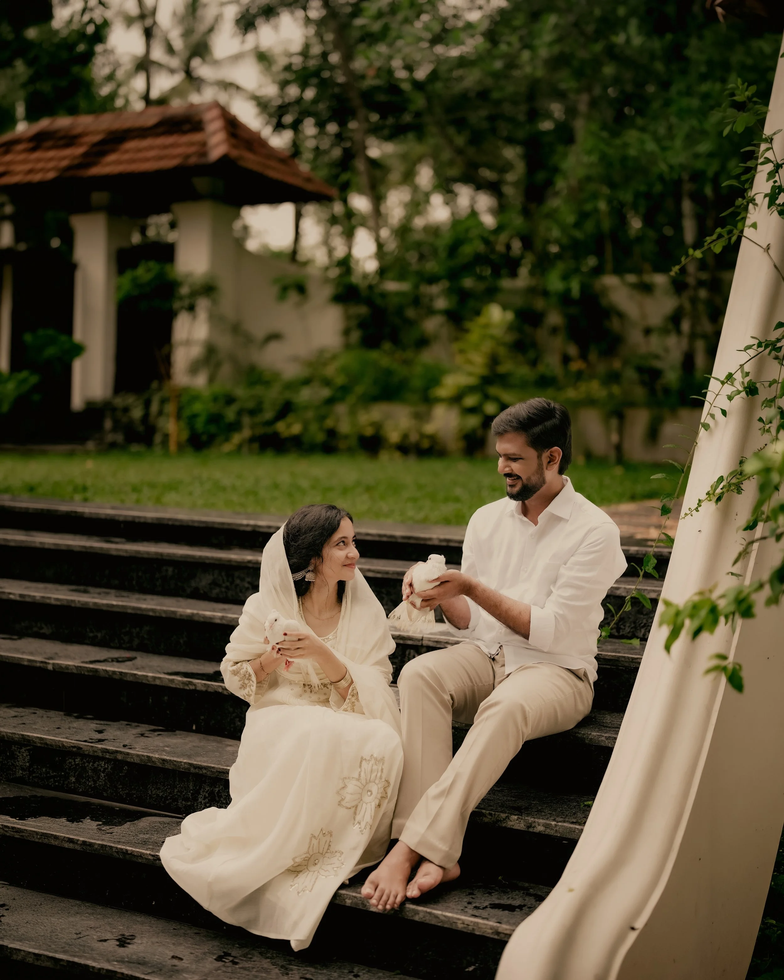 A man and a woman sitting on outdoor steps, exchanging gifts, with a white fabric draped nearby, in a garden setting with trees and a small building in the background.