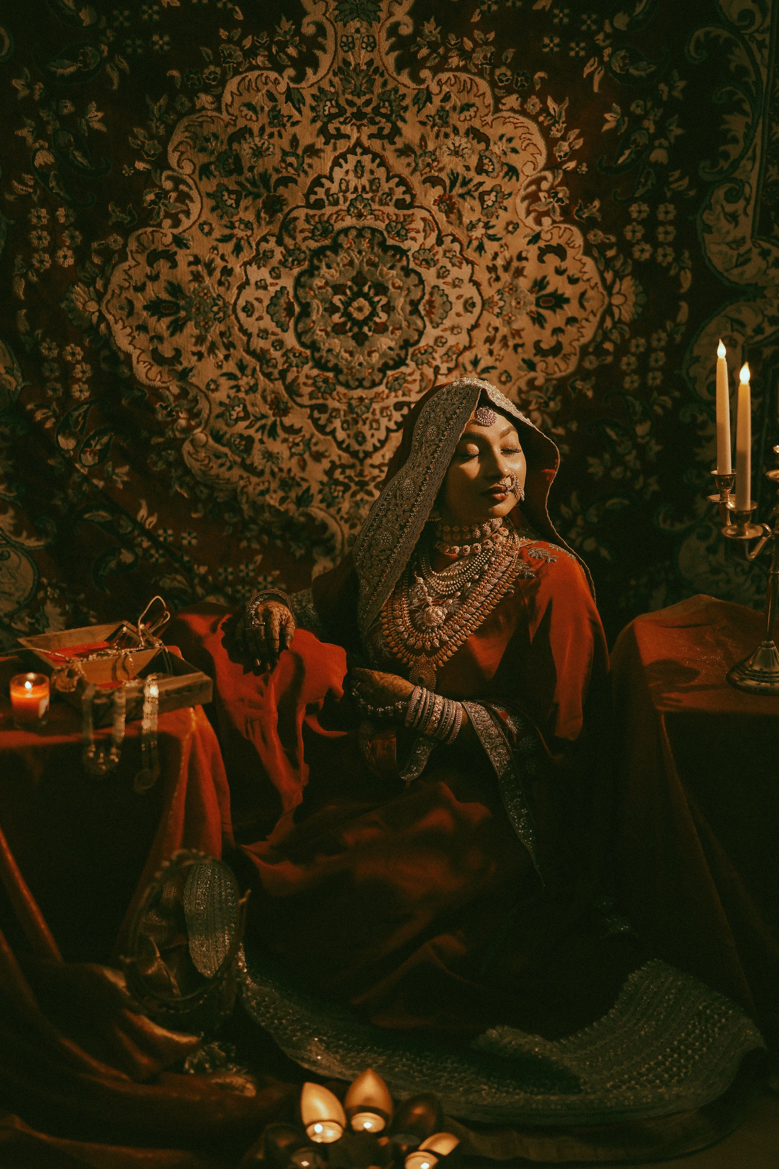 A woman dressed in traditional South Asian attire, wearing jewelry, sitting in front of an ornate Persian-style rug, with candles on her right, and various jewelry pieces on the table in front of her.