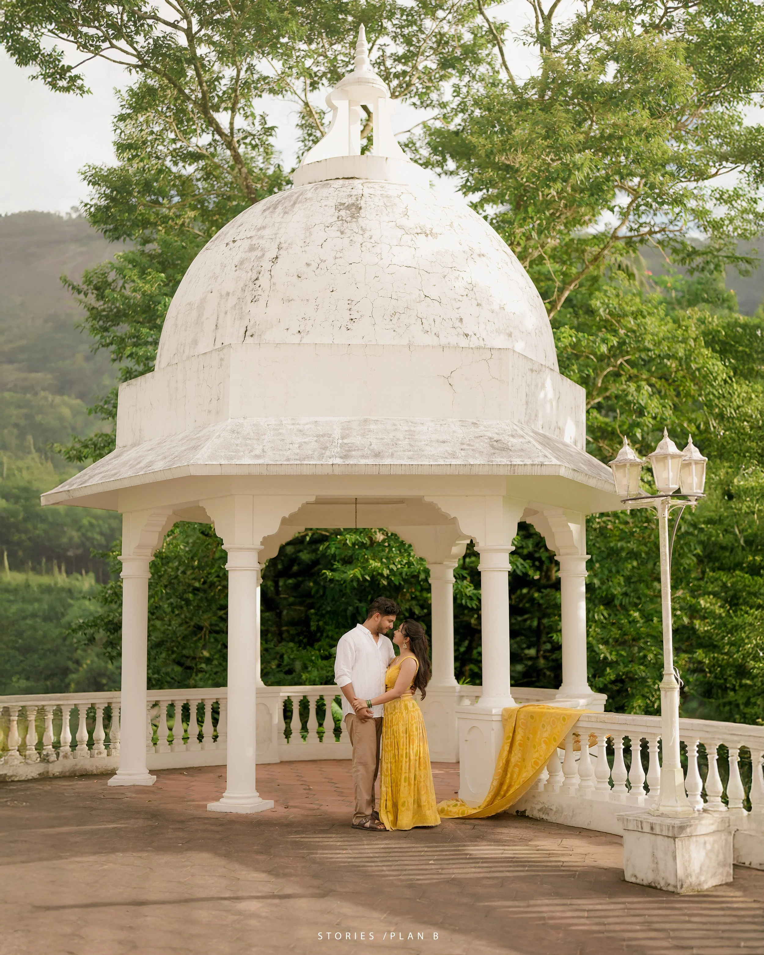 A man and woman stand close under a white gazebo, gazing at each other. The woman wears a long yellow dress, and the man wears a white shirt and beige pants. The setting is outdoors with green trees in the background. A yellow drape is tied to the side of the gazebo. The words 'STORIES/PLAN B' are at the bottom of the image.