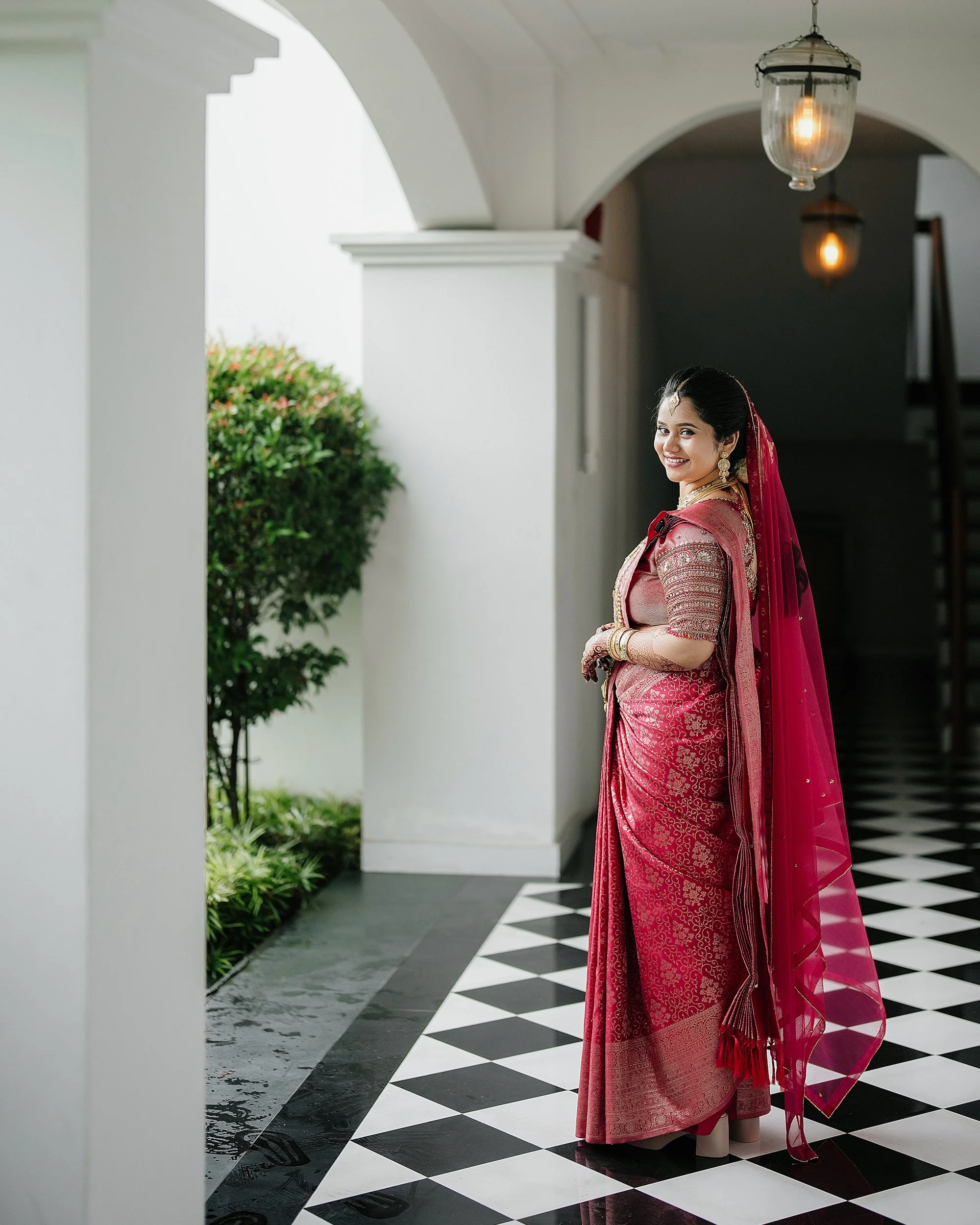 A woman in traditional Indian attire, including a pink saree and jewelry, stands in a hallway with black and white checkered floor tiles, white walls, and hanging lights. She is smiling and looking at the camera.