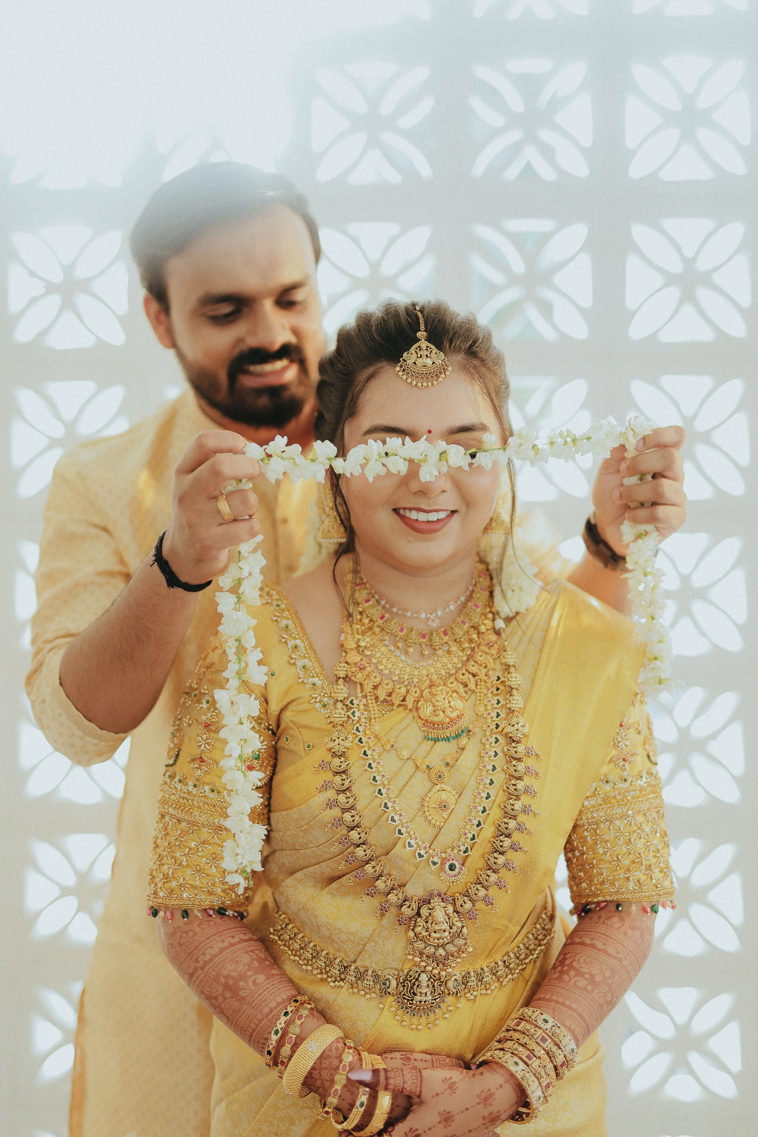 A Hindu wedding ceremony where a woman dressed in a traditional gold-colored saree, wearing ornate jewelry including necklaces, earrings, bangles, and a head ornament, smiles as a man in a yellow kurta ties a flower garland around her neck. The background features a light-colored decorative wall with cut-out patterns.