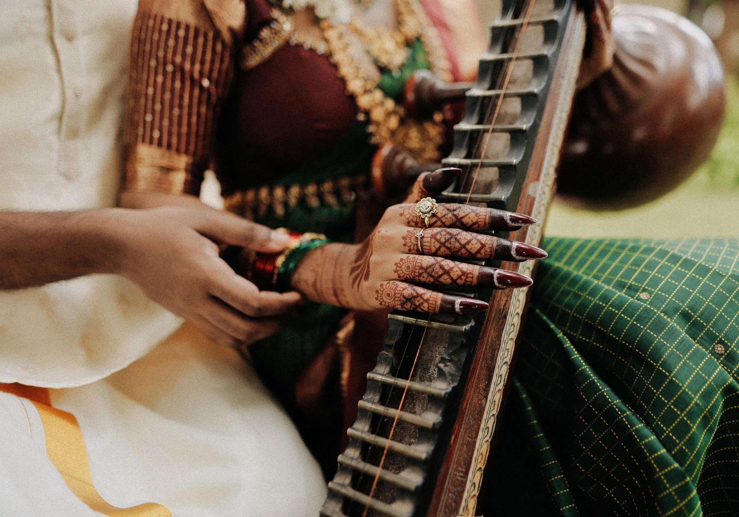 Close-up of a hand with henna designs and rings playing a traditional musical instrument, with a person wearing a green and black checkered garment nearby.