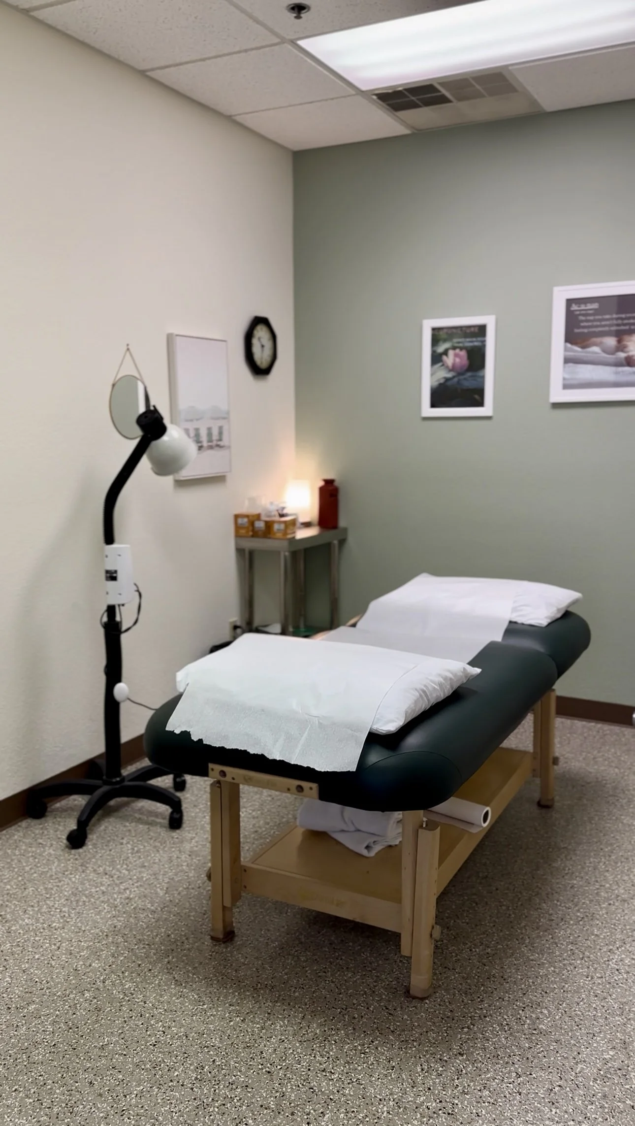 Medical treatment room with an examination table covered with paper sheets, a lamp, and framed pictures on the wall.