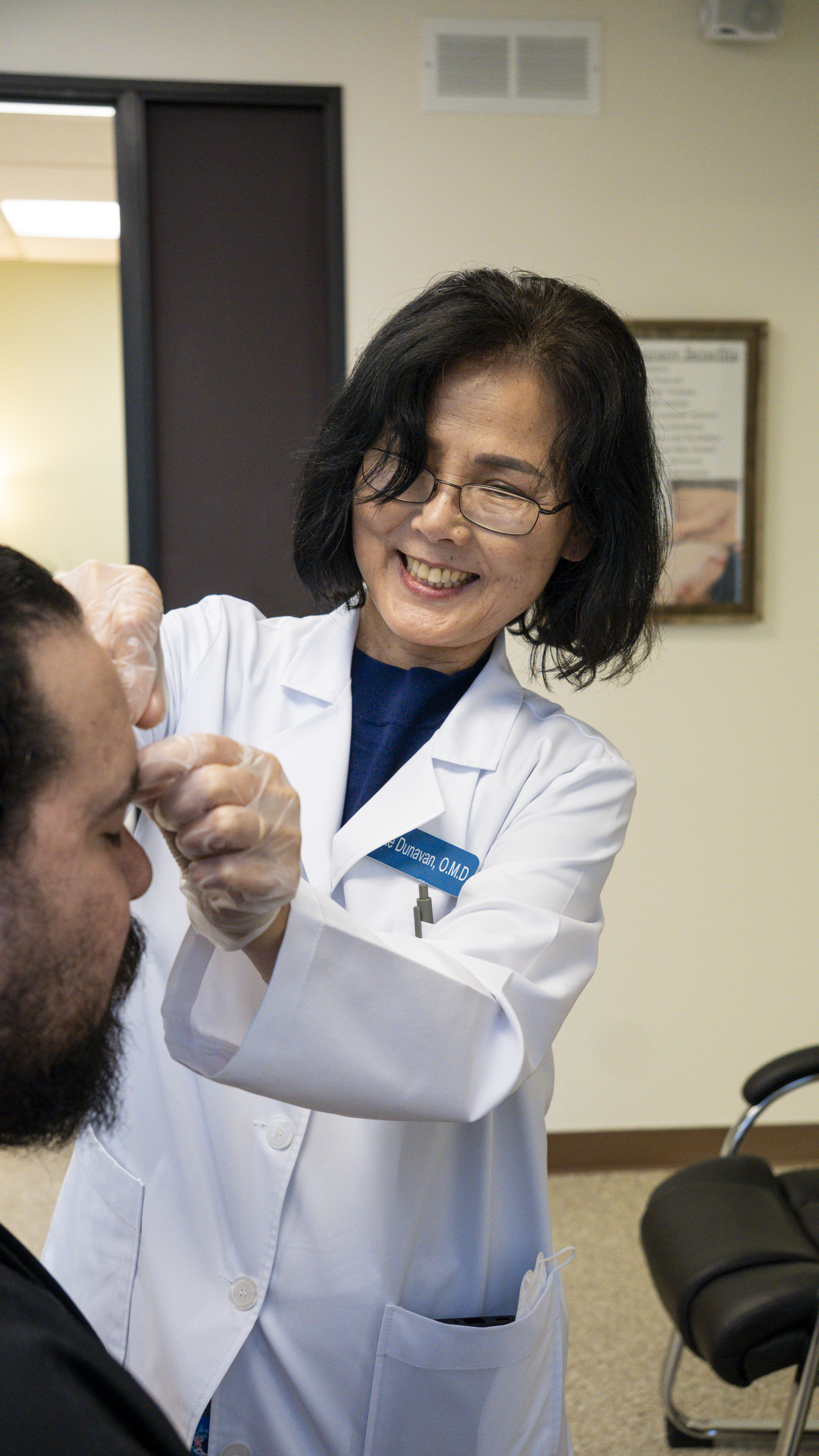 A female healthcare professional with glasses and shoulder-length black hair, smiling as she administers a medical procedure to a male patient in a clinic setting.