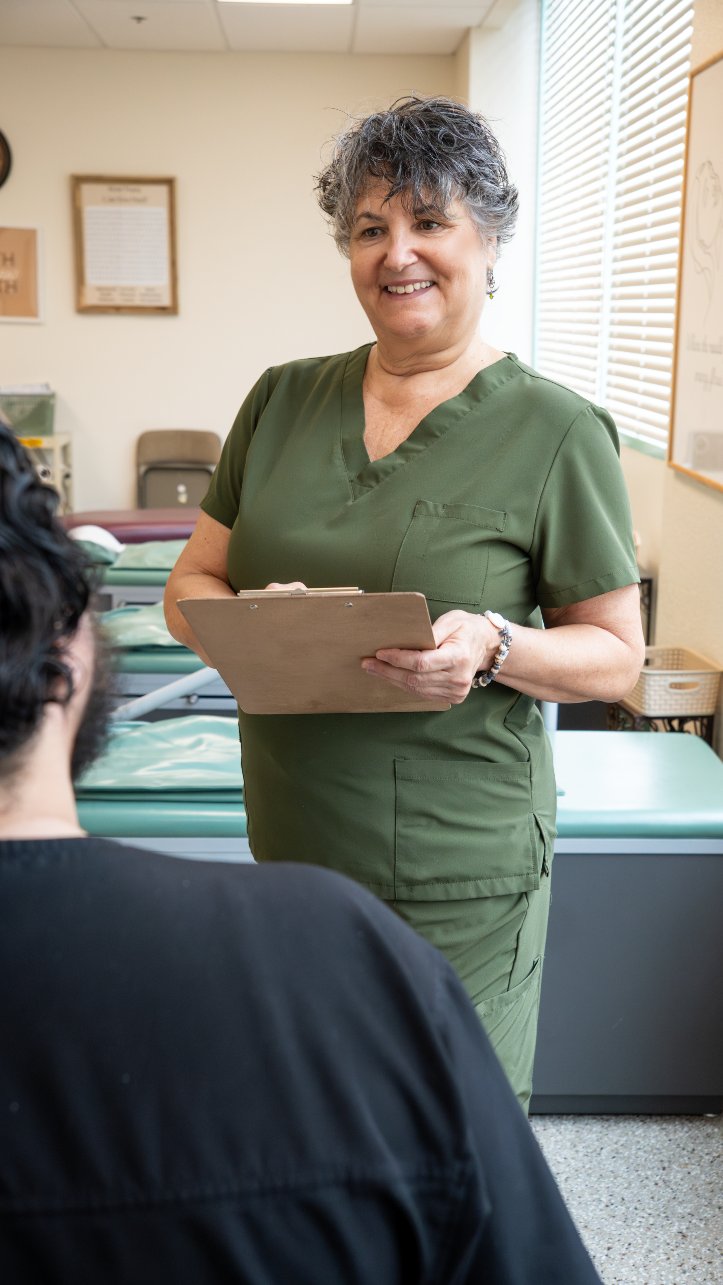 A middle-aged woman with curly gray hair and earrings wearing green medical scrubs, holding a clipboard, smiling and talking to a person with dark hair, seated in a healthcare setting with a bed and medical equipment in the background.