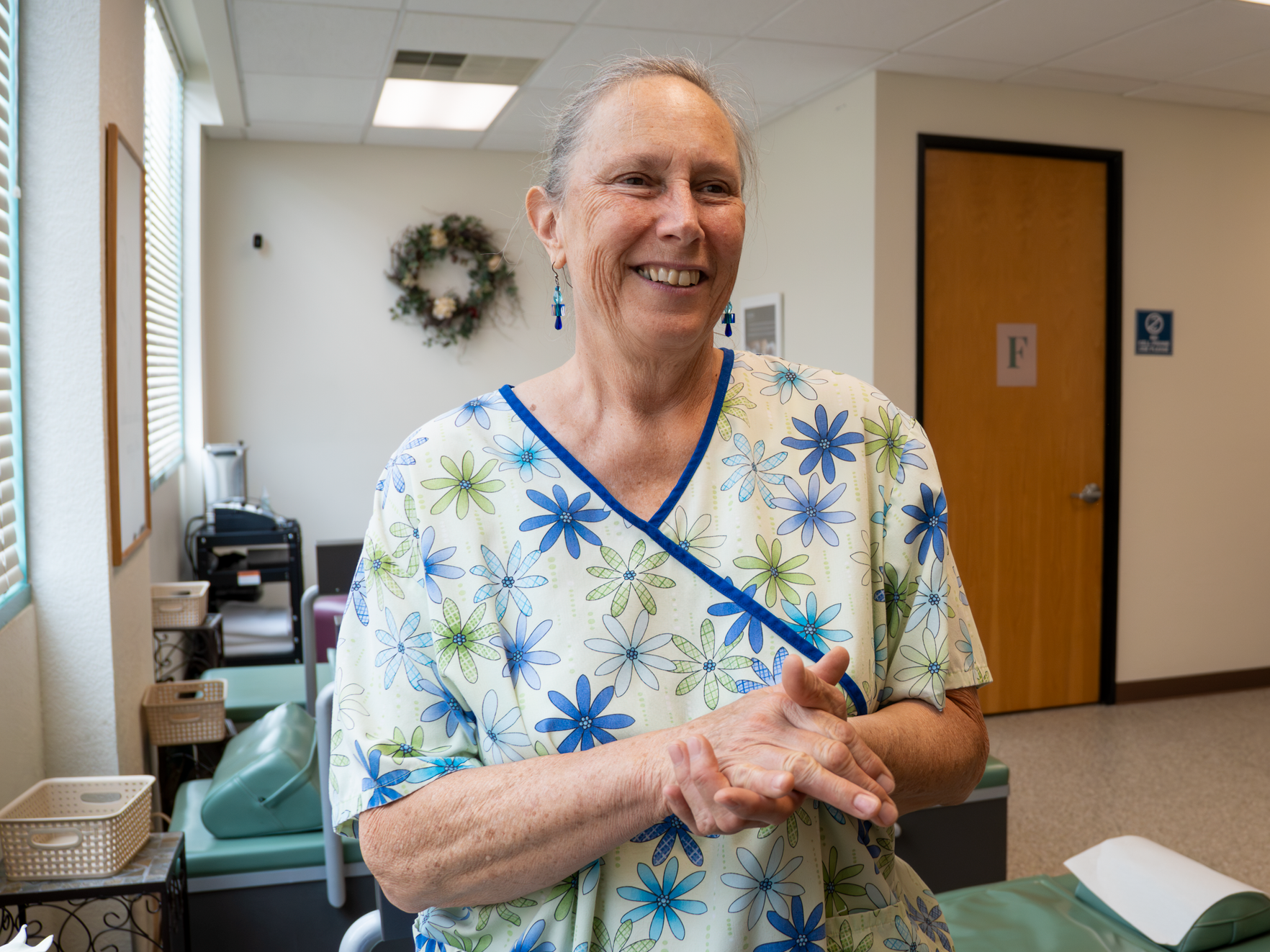 An elderly woman in a floral medical gown smiling indoors in a clinical setting.
