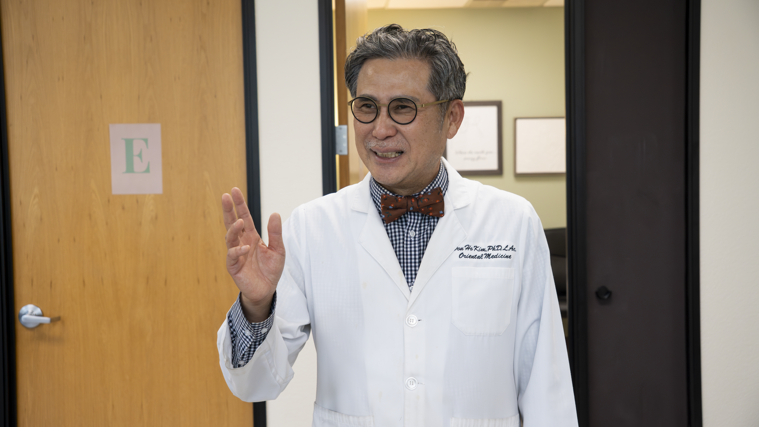 A doctor with glasses and a bowtie waving while standing in a doorway. The doctor is wearing a white coat with a name tag that reads 'Jon Ho Kim, PhD, L.Ac. Oriental Medicine.' The background shows a hallway with framed pictures on the wall.