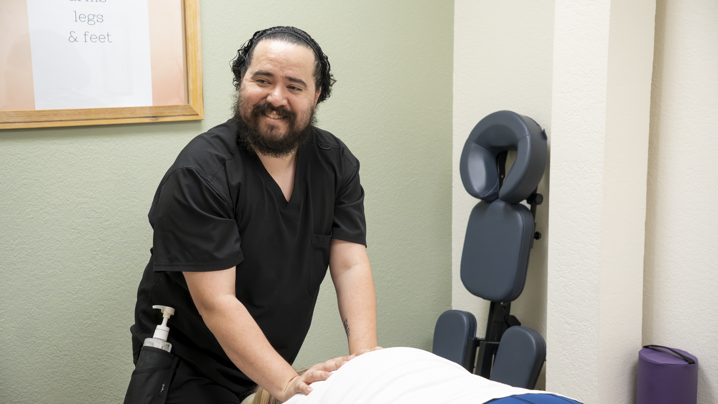 A man with a beard and long hair tied back, wearing black scrubs, providing a massage to a person lying face down on a massage table in a wellness or physical therapy clinic.