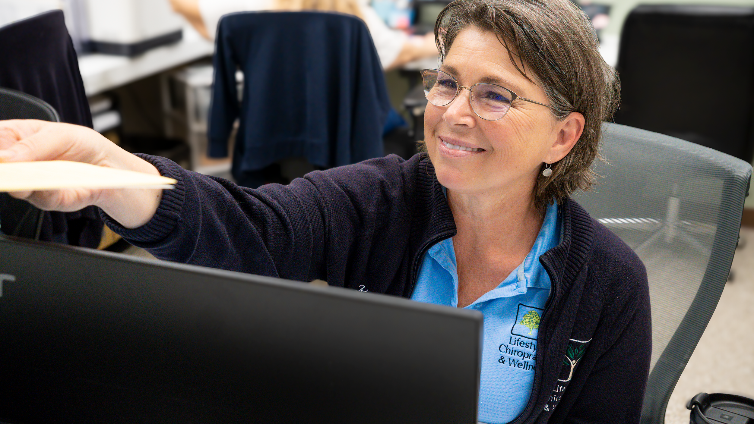 A smiling woman with short brown hair, glasses, and earrings, wearing a blue polo shirt with a logo, is reaching out to accept a paper or document in an office or classroom setting with several chairs and desks.