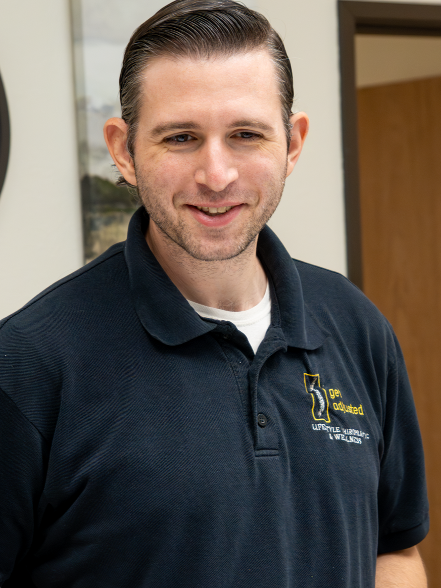 A man with slicked-back hair and a beard wearing a navy blue polo shirt with a logo that reads "get adjusted" and additional text, standing indoors with a neutral background.