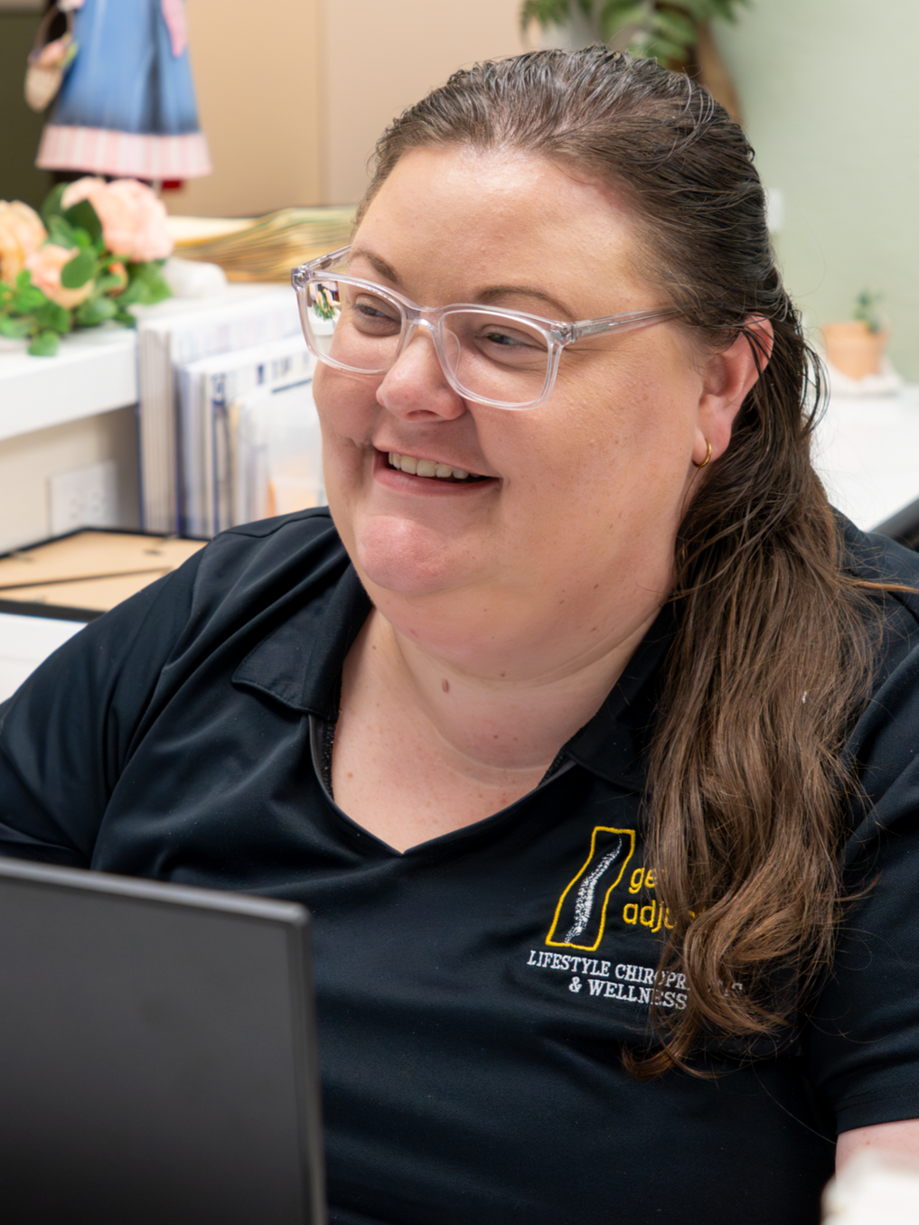 A woman with glasses smiling while working on a computer in an office environment.
