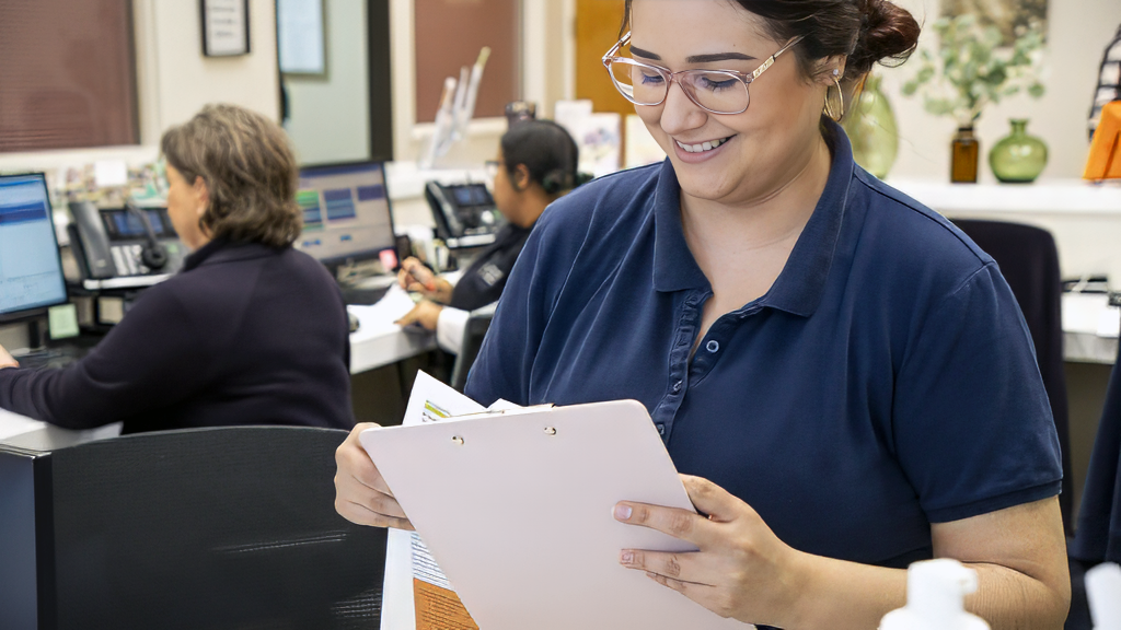 A smiling woman in glasses and a navy polo shirt holding a clipboard in an office with multiple computer workstations and colleagues in the background.