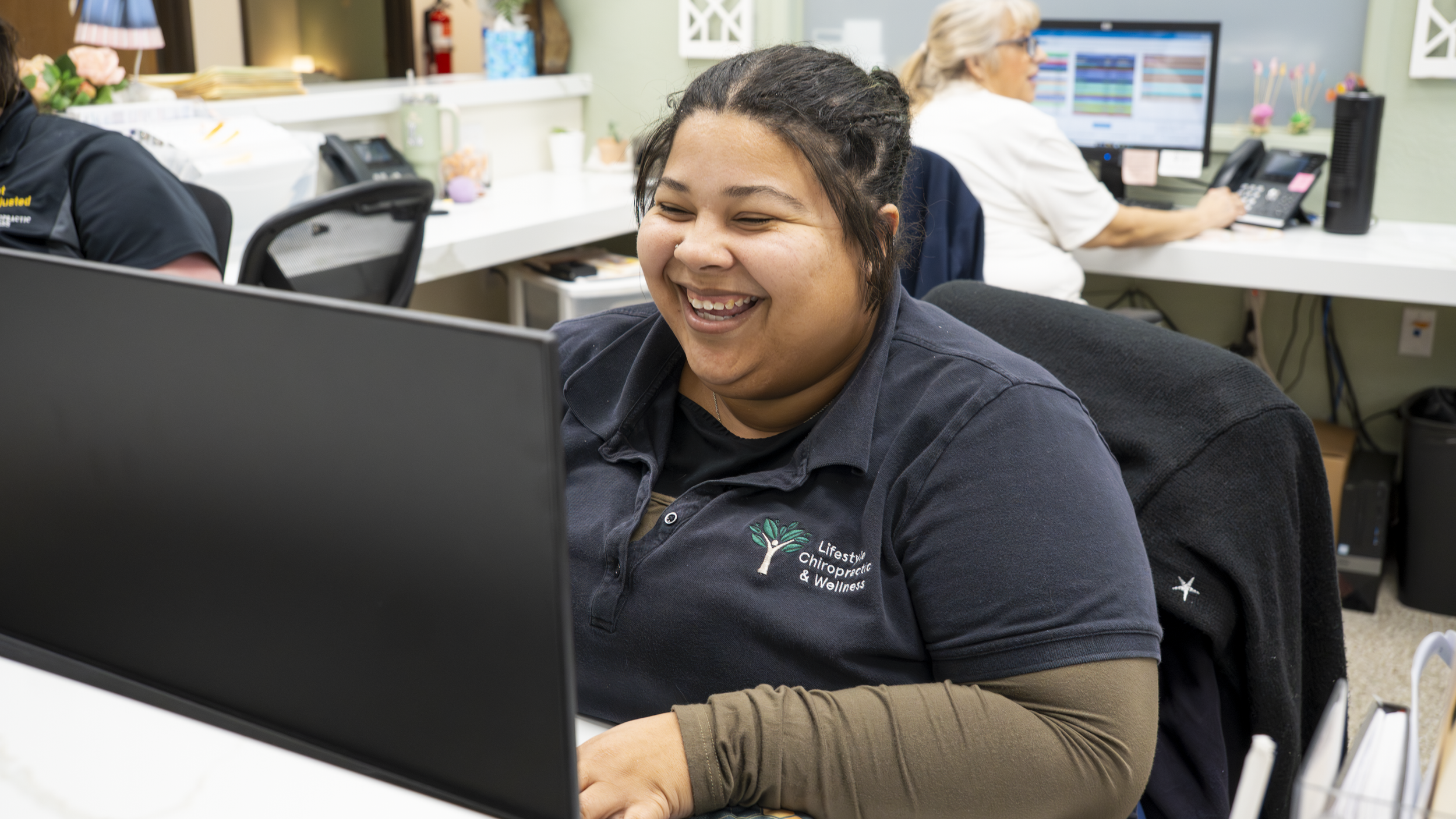Joyful woman working at a computer in an office, with other people working in the background.