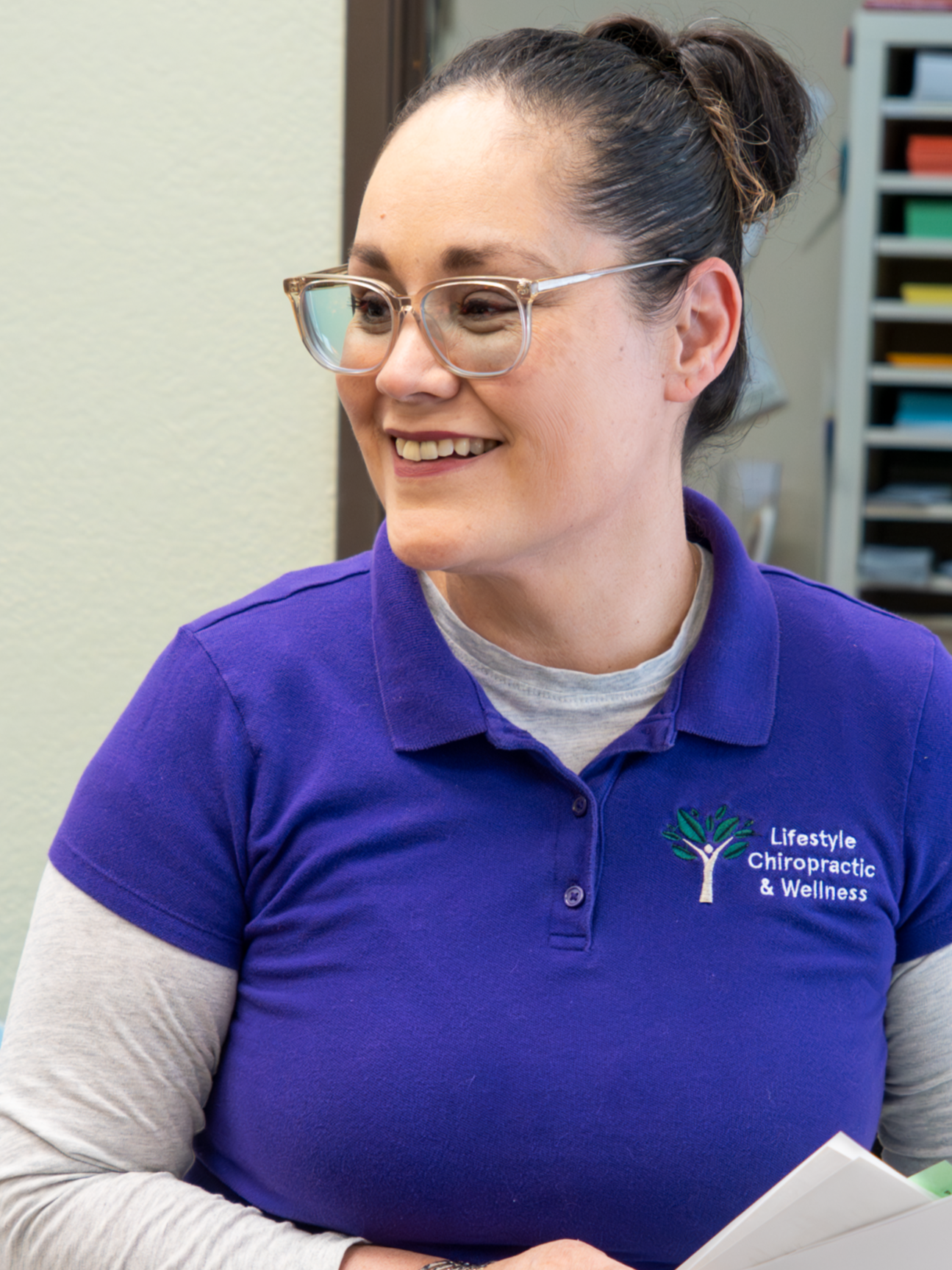 Smiling woman wearing glasses and a purple polo shirt with a logo for Lifestyle Chiropractic & Wellness, holding papers in her hands, standing in an office environment.
