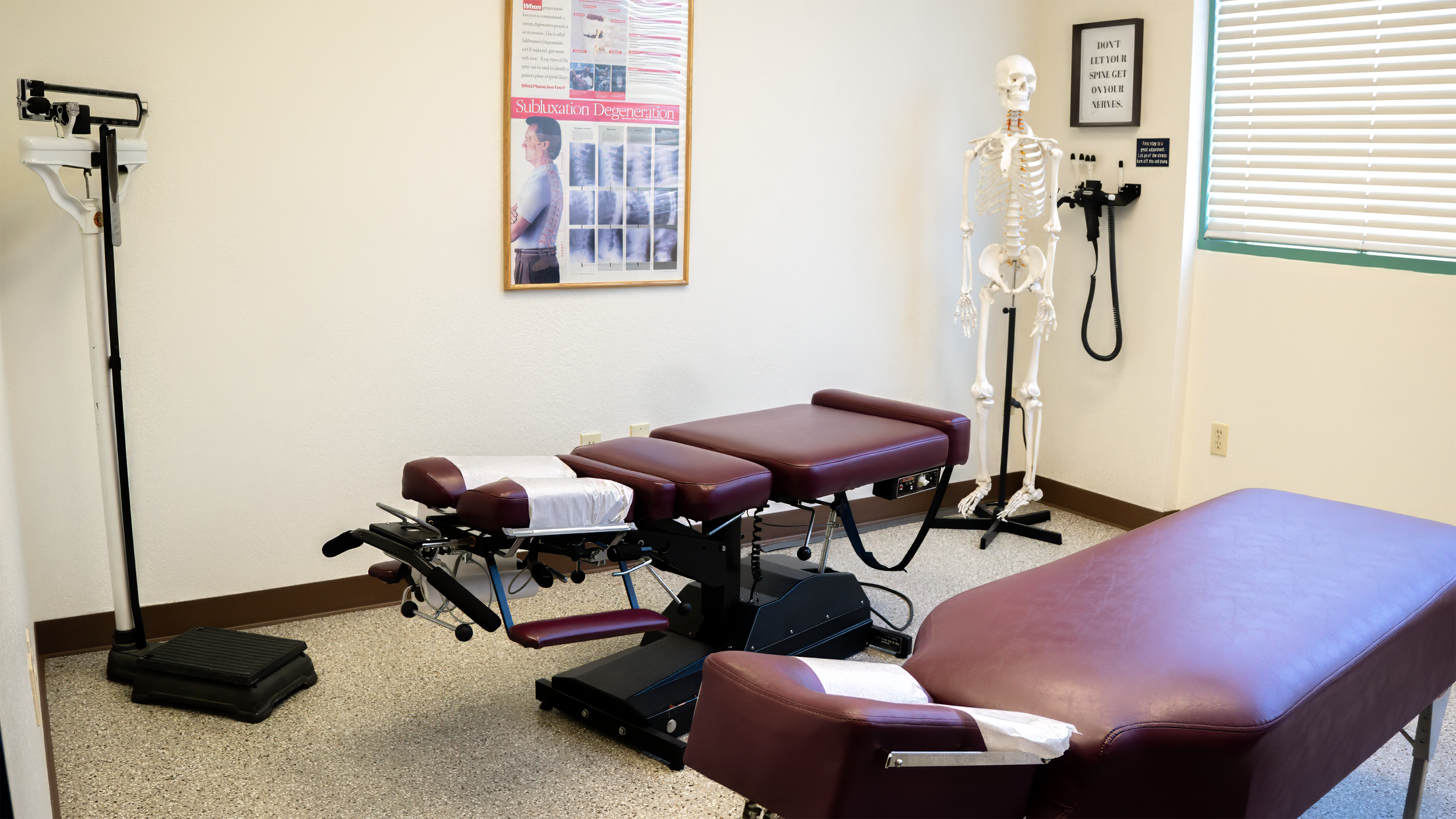 Exam room with chiropractic treatment table, skeleton model, medical posters, and equipment, including a blood pressure cuff and footstep analyzer