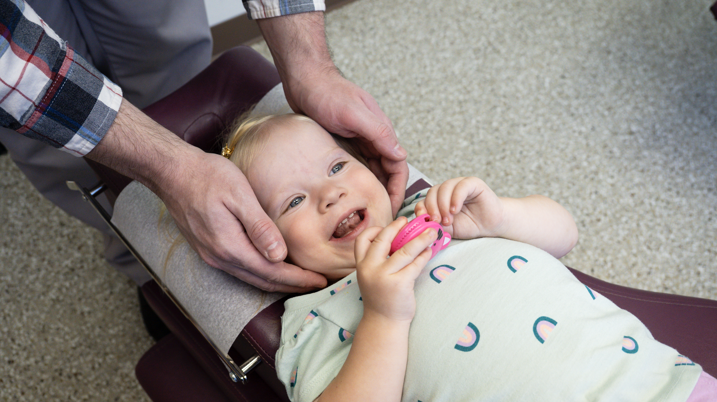 A young girl with blonde hair lying on a medical examination table, wearing a light-colored shirt with rainbows, holding a pink toy, and being gently held by a person wearing a plaid shirt with rolled sleeves, who is supporting her head and shoulders with their hands.