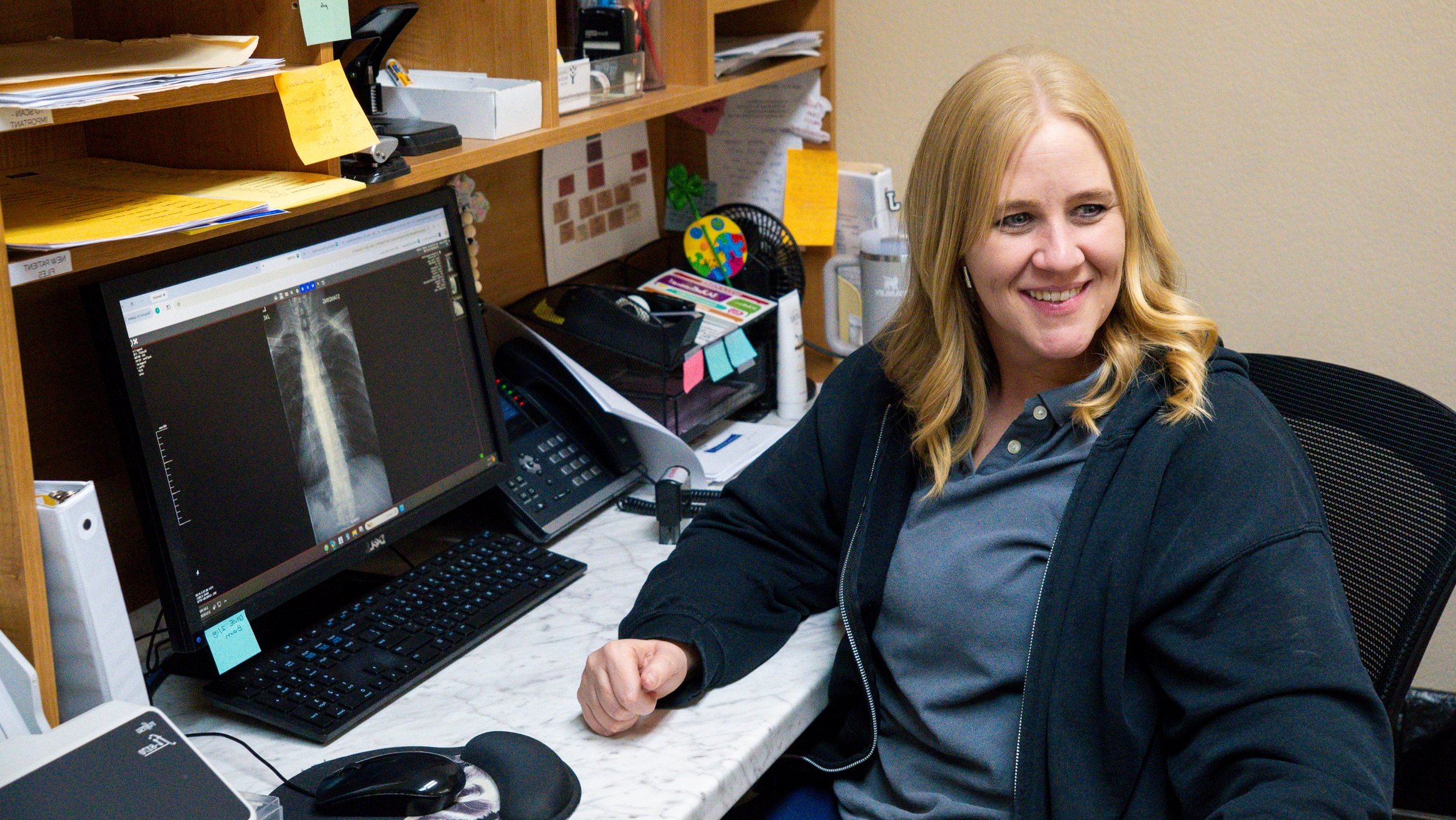A woman with long blonde hair smiling, sitting at a cluttered desk with a computer showing a medical x-ray image, surrounded by office supplies and papers.