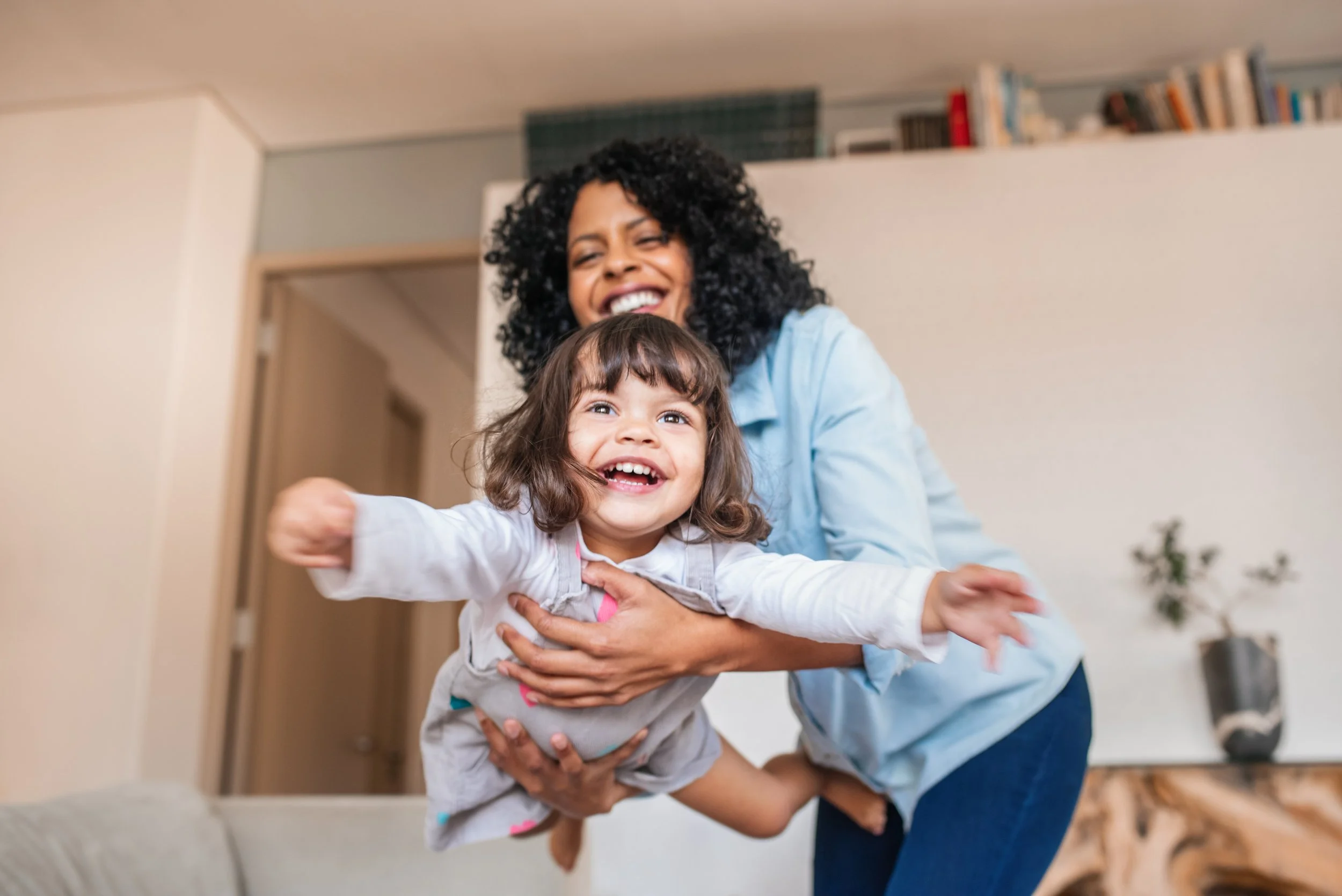 A woman with curly black hair smiling and holding a happy young girl with brown hair, who is pointing towards the camera in a living room.