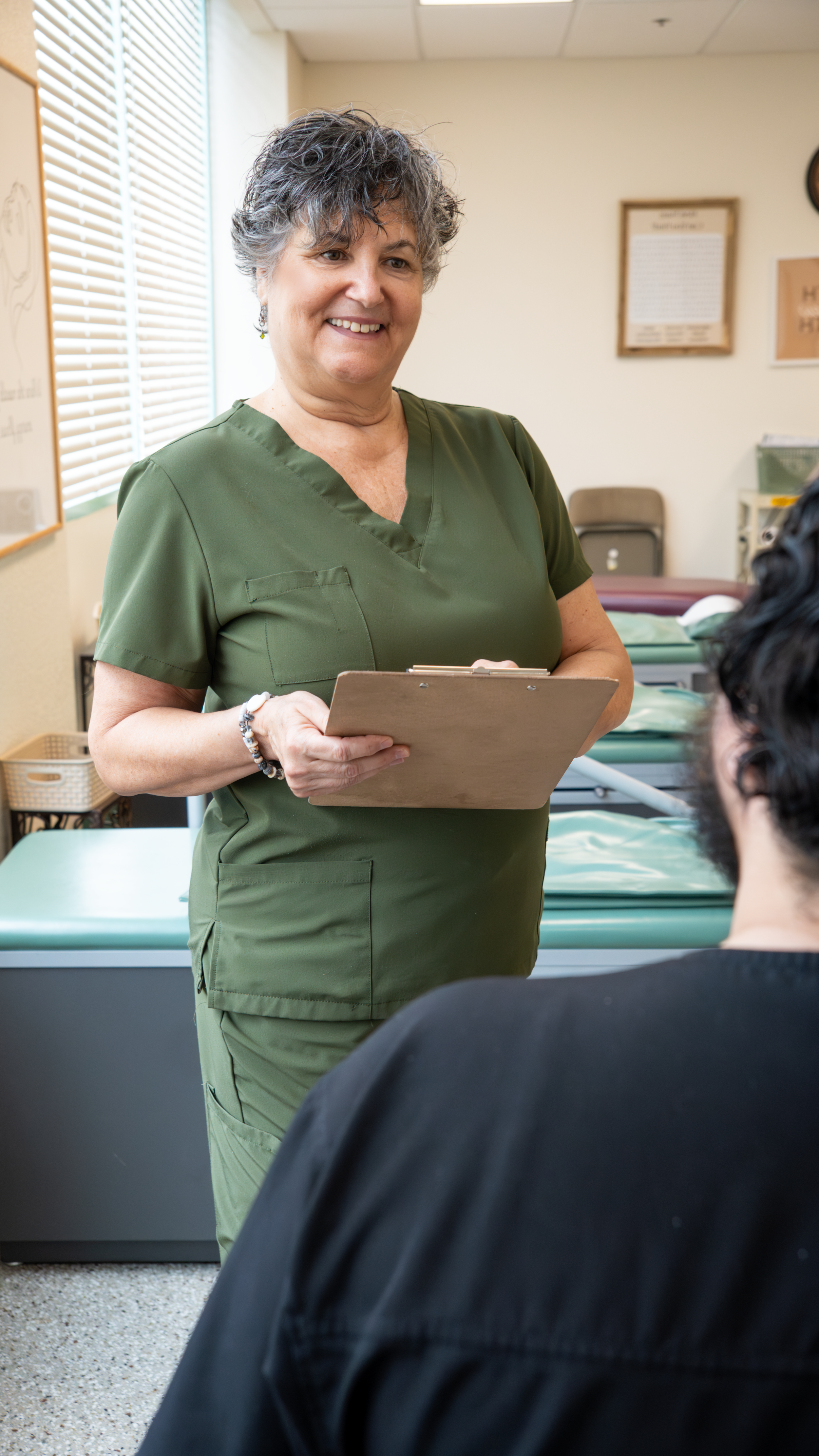 A healthcare worker in green scrubs smiling while holding a clipboard and talking to a patient in a clinic or medical office setting.